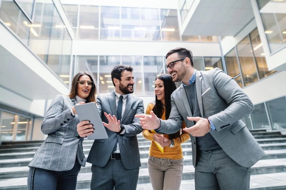 Group of four business professionals smiling and interacting with a tablet in a modern office building lobby.