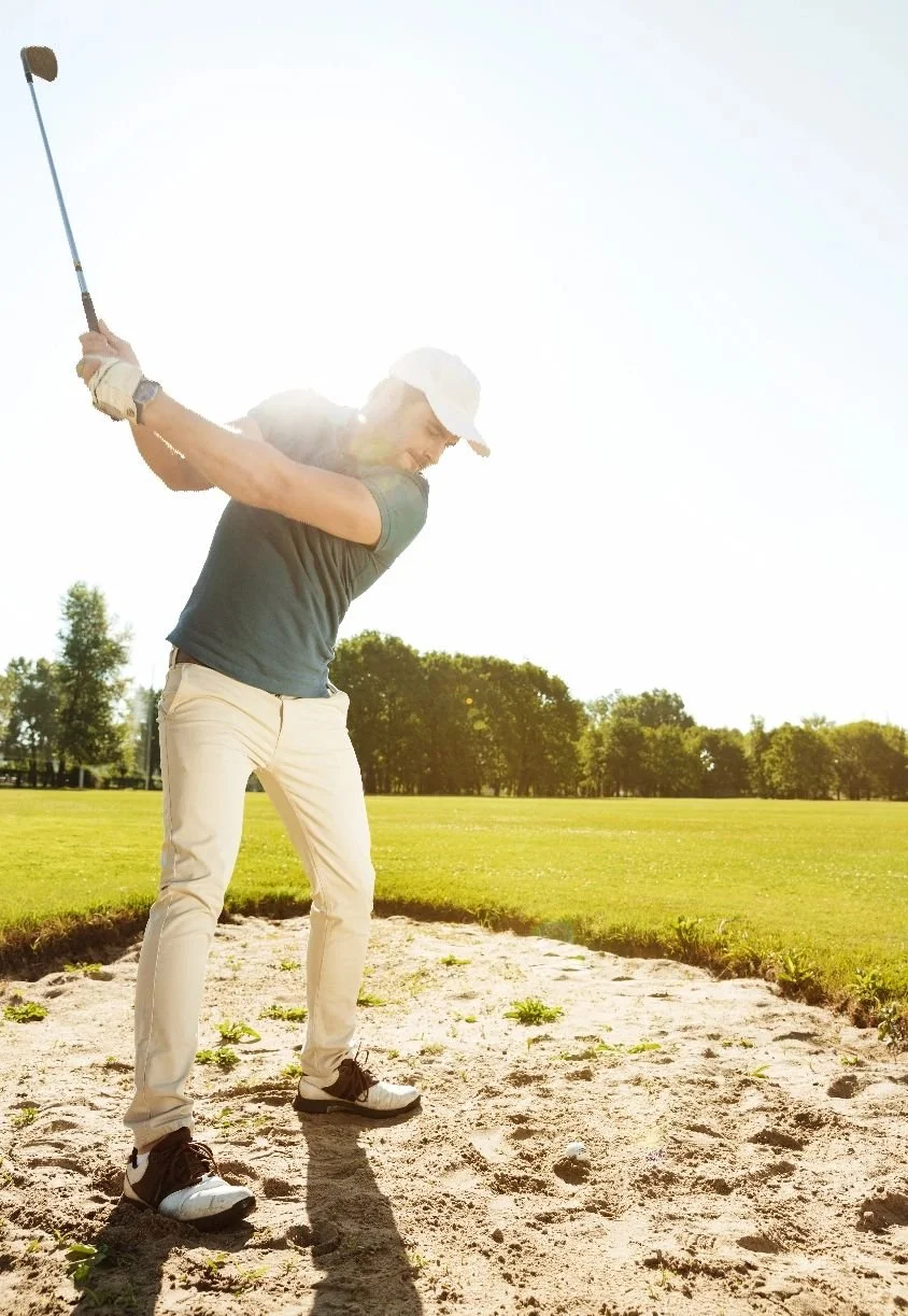 Person swinging a golf club on a golf course in bright, sunny weather.