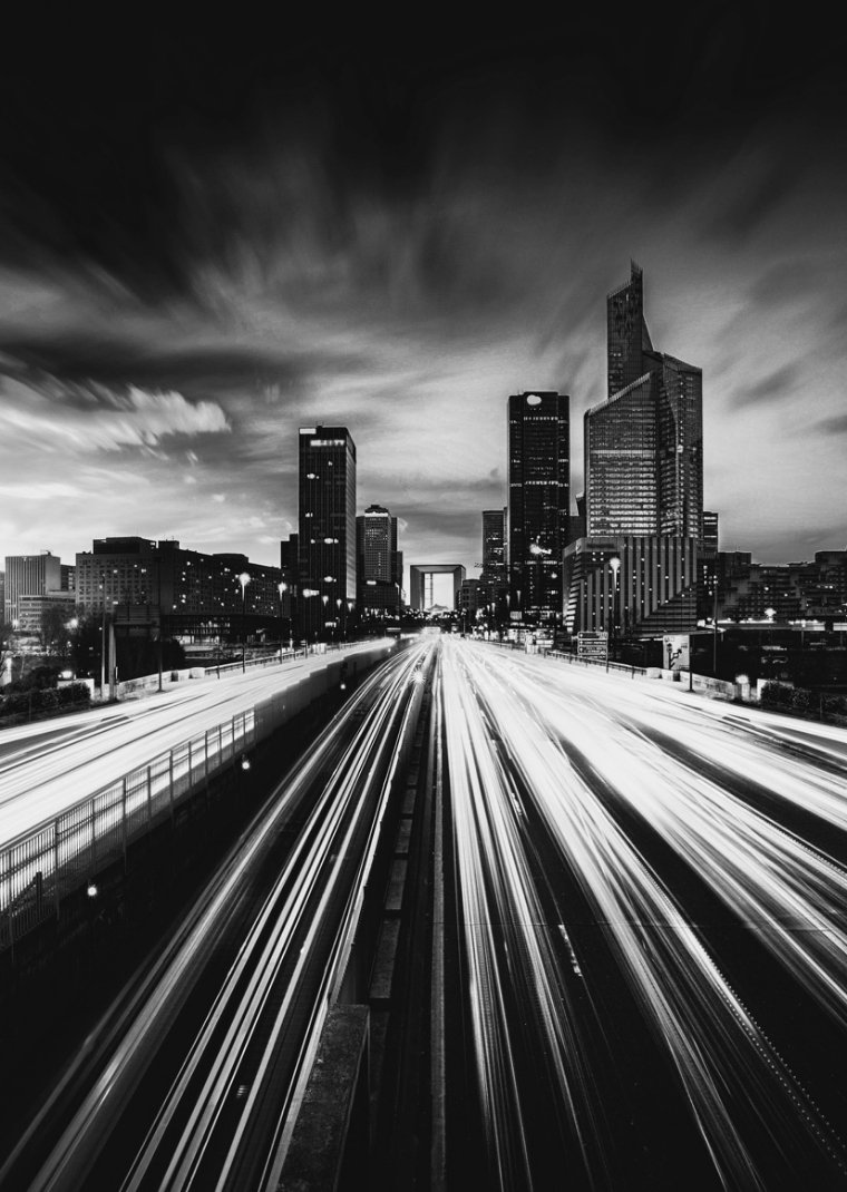 Black and white cityscape featuring tall modern skyscrapers with streaks of light from moving vehicles on a busy highway in the foreground.