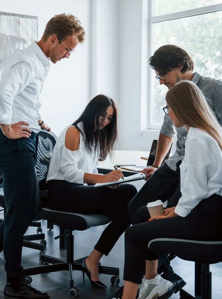 Four young professionals in an office discussing around a seated woman who is taking notes