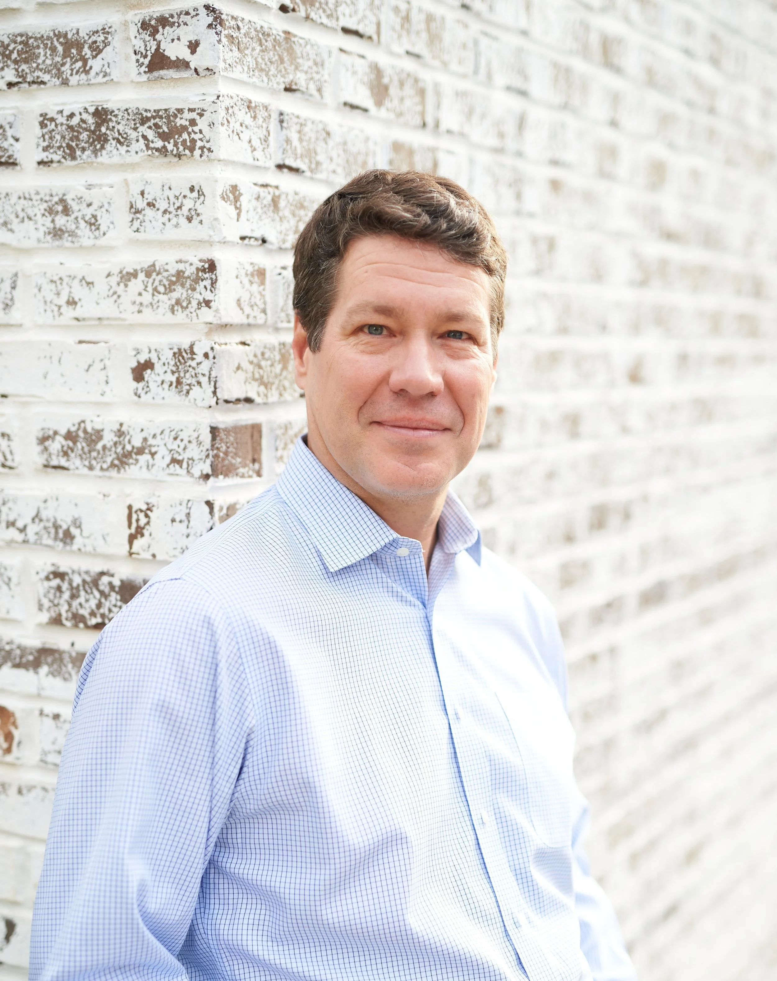A man with brown hair, wearing a light blue checkered shirt, standing against a white brick wall outside.