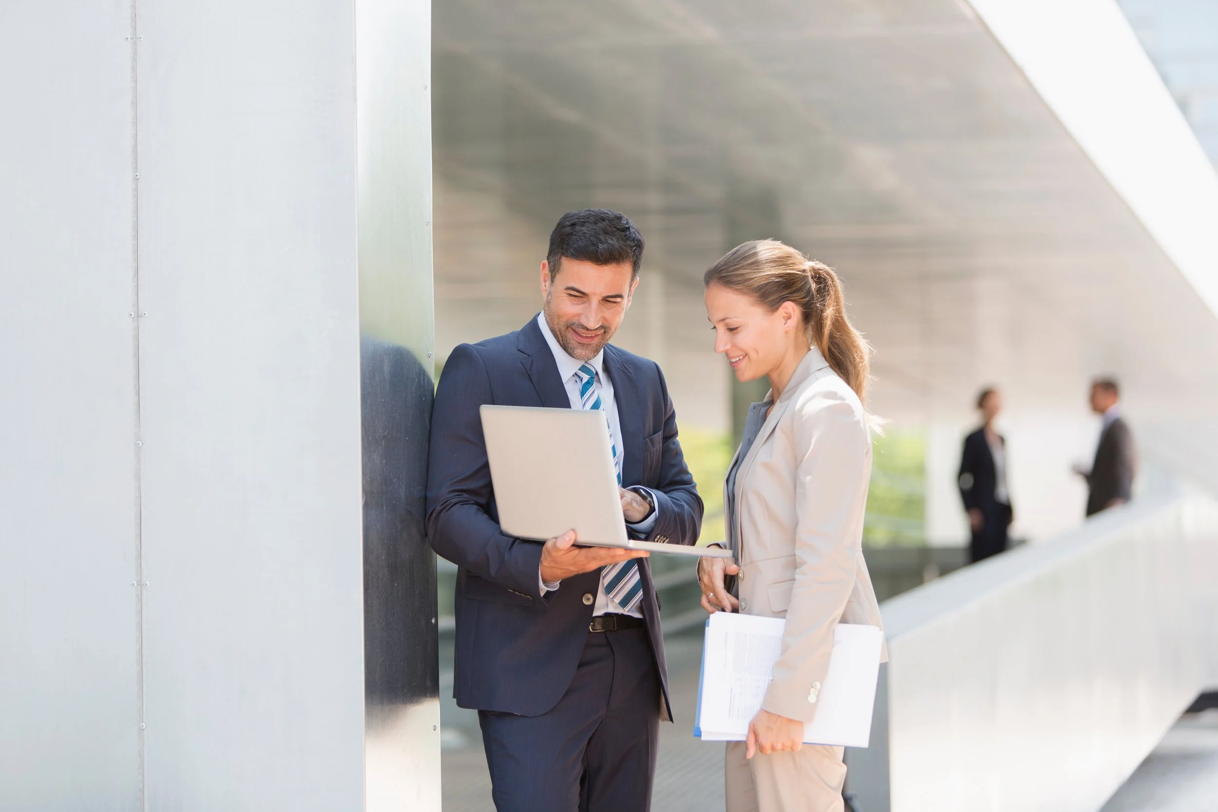 Two businesspeople, a man and a woman, are standing outdoors in business attire, looking at a laptop and smiling. The woman is holding a clipboard and some papers, and they appear to be engaged in a work-related discussion.