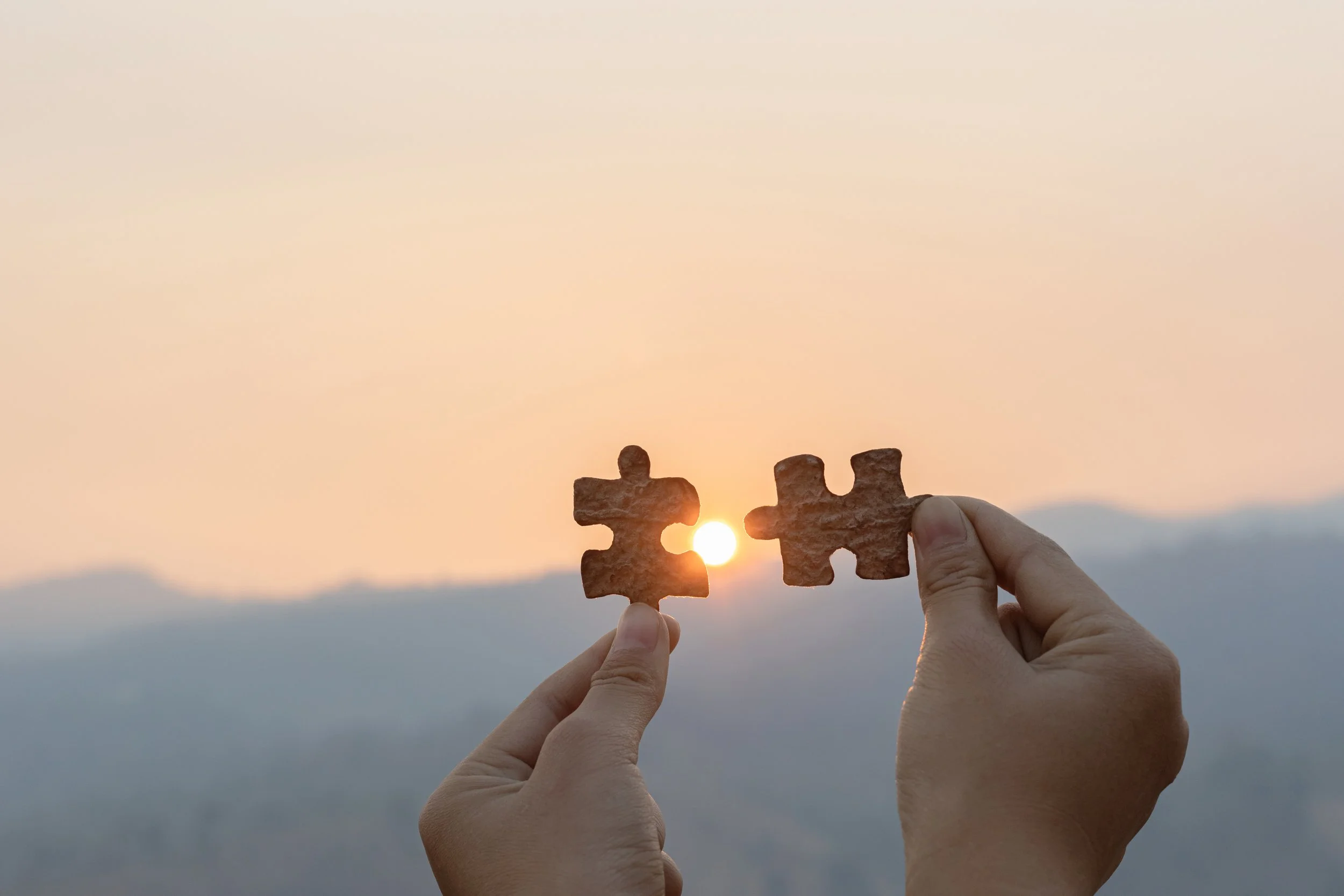 Two hands holding puzzle pieces in front of a sunset with mountains in the background, with the sun aligning between the puzzle pieces.
