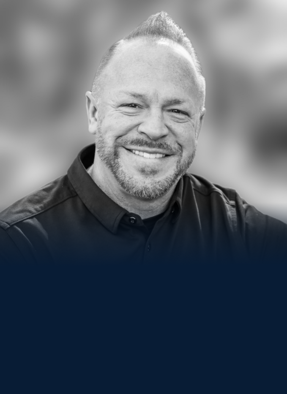 Black and white portrait of a smiling man with short hair, beard, wearing a dark button-up shirt, outdoors.