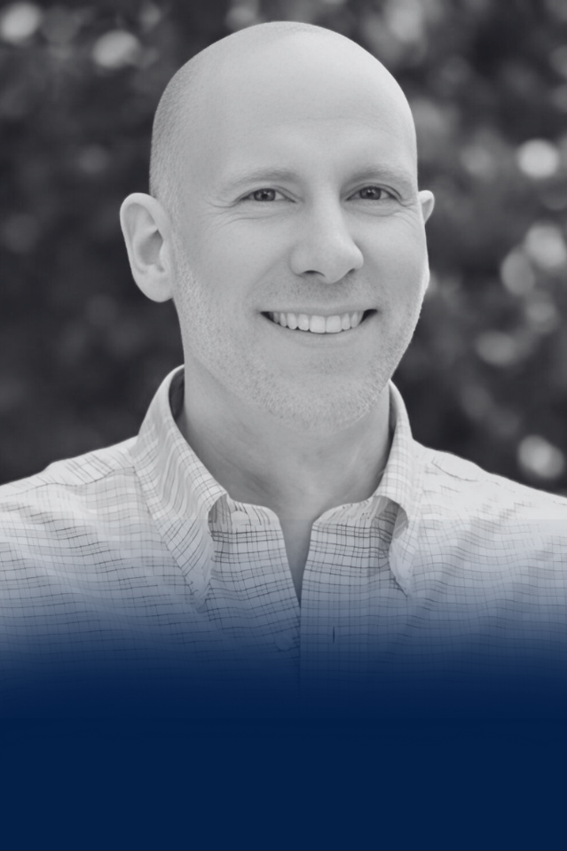 Black and white portrait of a bald man with a broad smile, wearing a plaid button-up shirt, standing outdoors with a blurred natural background.