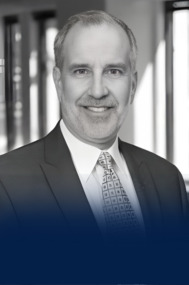 A smiling middle-aged man with gray hair, wearing a suit, tie, and white shirt, standing in an office with large windows in the background.