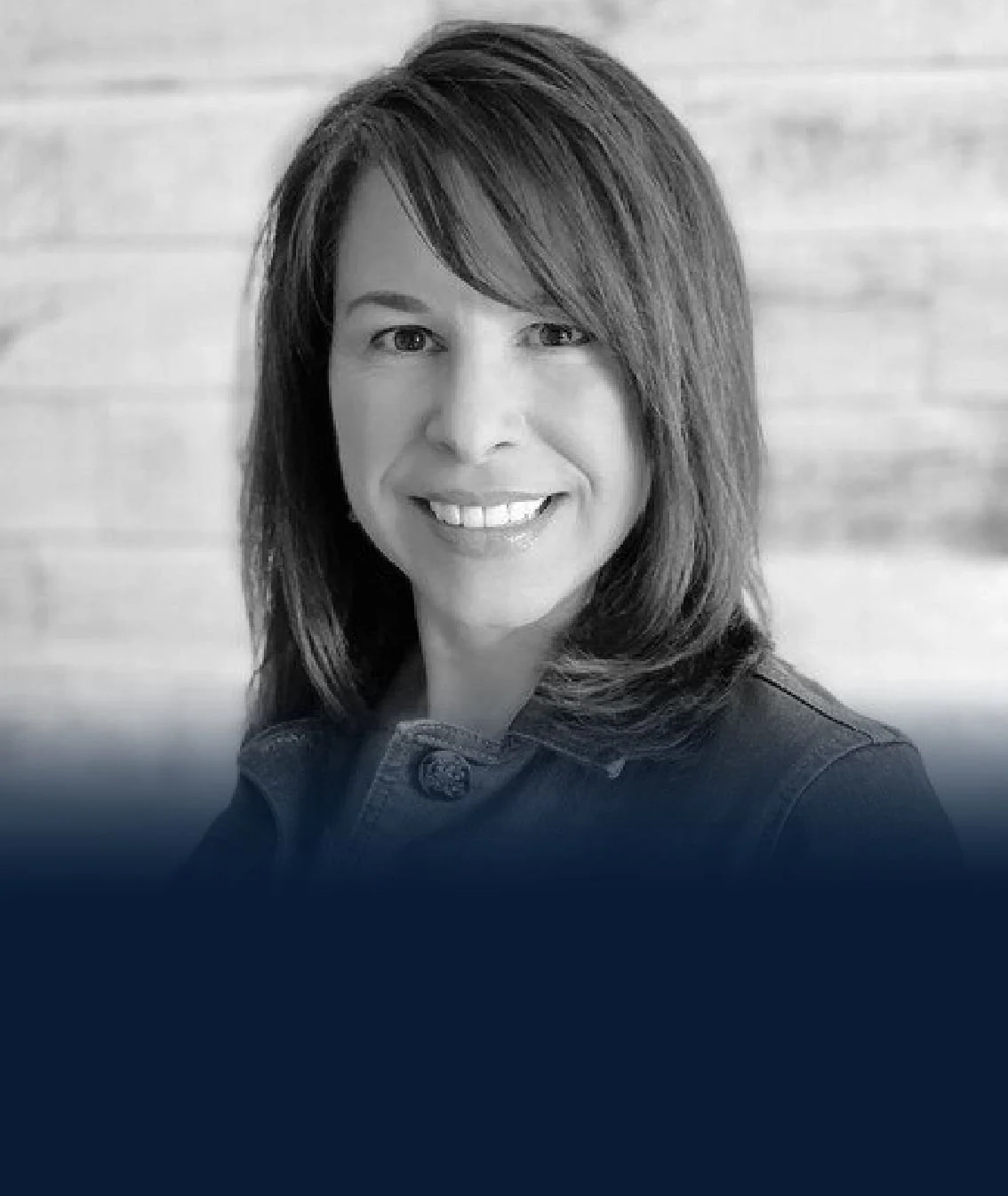 Black and white portrait of a woman with shoulder-length hair smiling at the camera, wearing a denim jacket, with a wooden background.