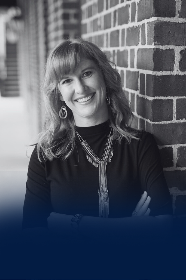Smiling woman with wavy hair, wearing earrings and a necklace, standing against a brick wall.
