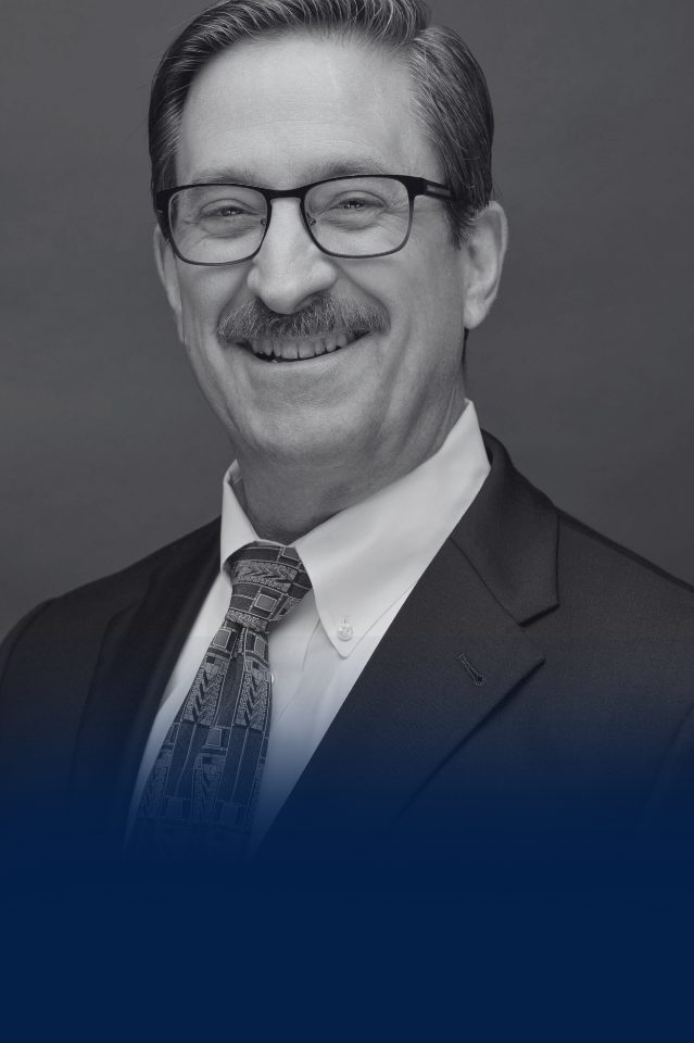 Black and white portrait of a smiling man with glasses, a mustache, wearing a dark suit, white shirt, and patterned tie.