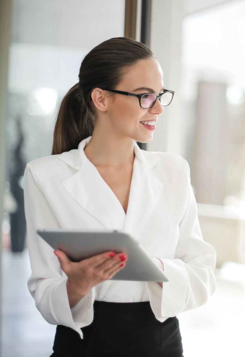 A woman with glasses and a white blazer holding a tablet in an office environment, smiling and looking to the side.