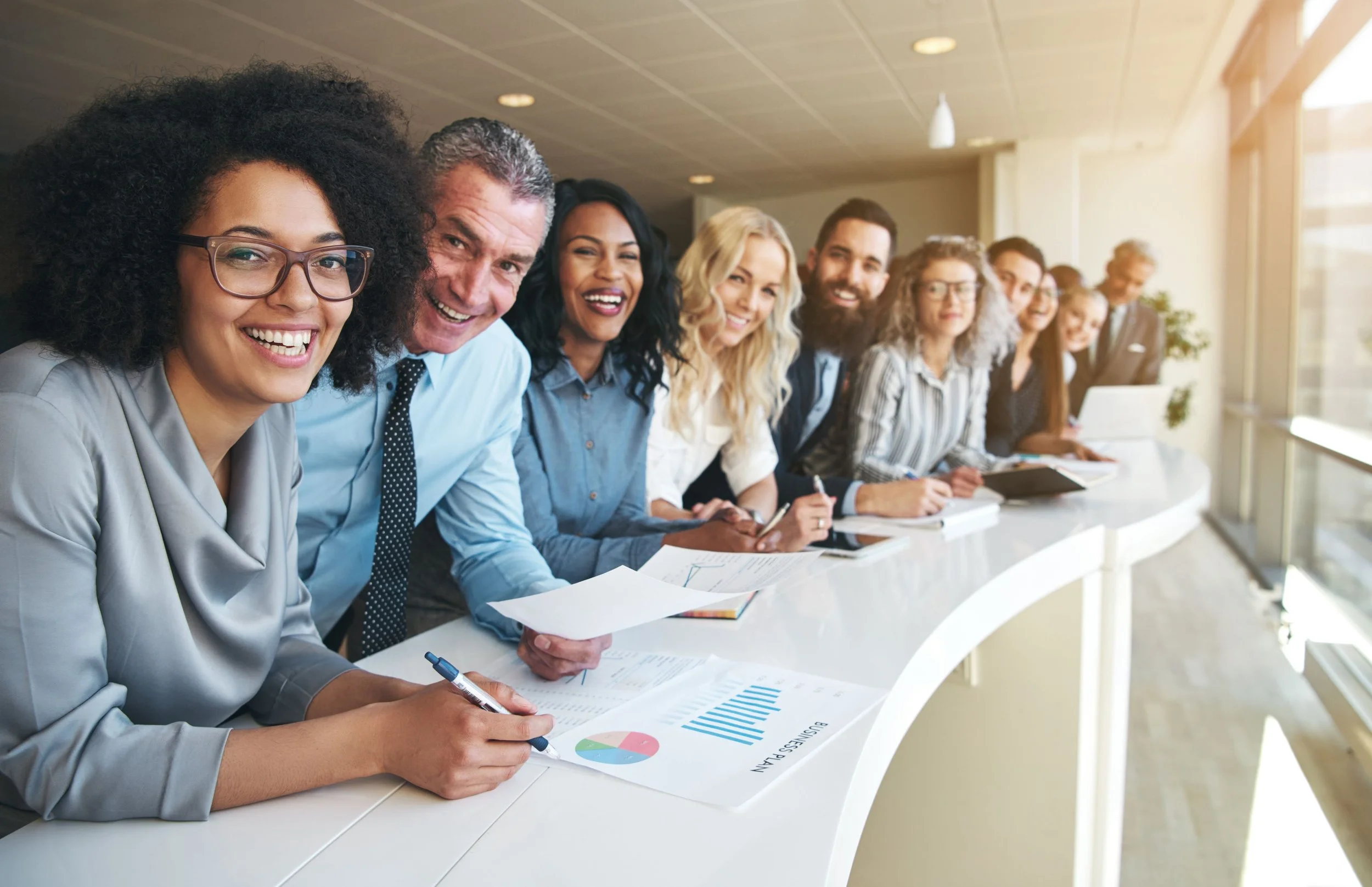 A diverse group of people smiling and sitting at a long conference table in a modern office, with documents and electronic devices in front of them, during a meeting or presentation.