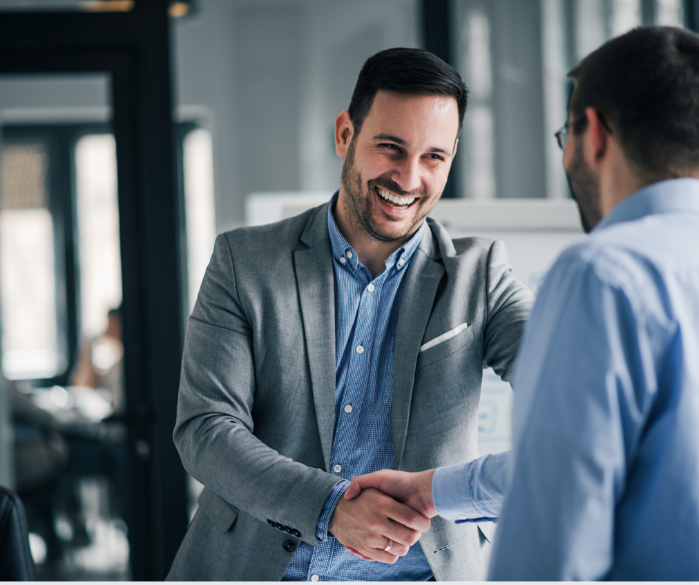 Two men in business attire shaking hands and smiling in a modern office setting.