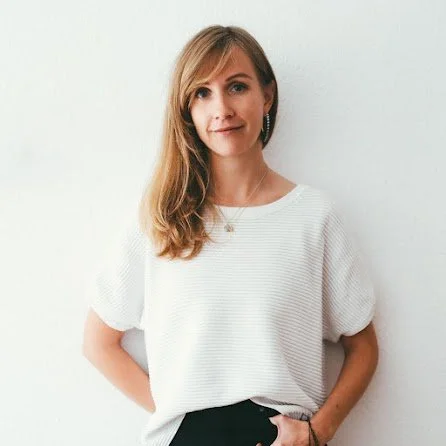 Young woman with long, wavy, light brown hair wearing a white textured short-sleeve blouse and black pants, standing against a plain white wall.