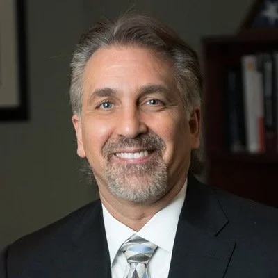 A professional man with gray hair and beard smiling in an office setting, dressed in a suit and tie.