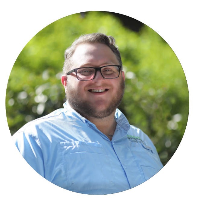 A smiling man with glasses and a beard wearing a light blue collared shirt outdoors with green trees in the background.