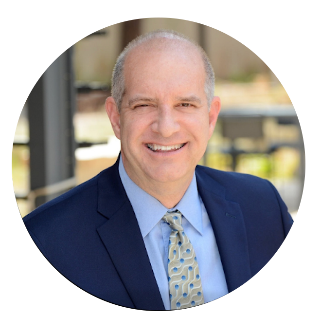 A smiling middle-aged man with short gray hair, wearing a dark blue suit, light blue shirt, and patterned tie, standing outdoors in a professional setting.