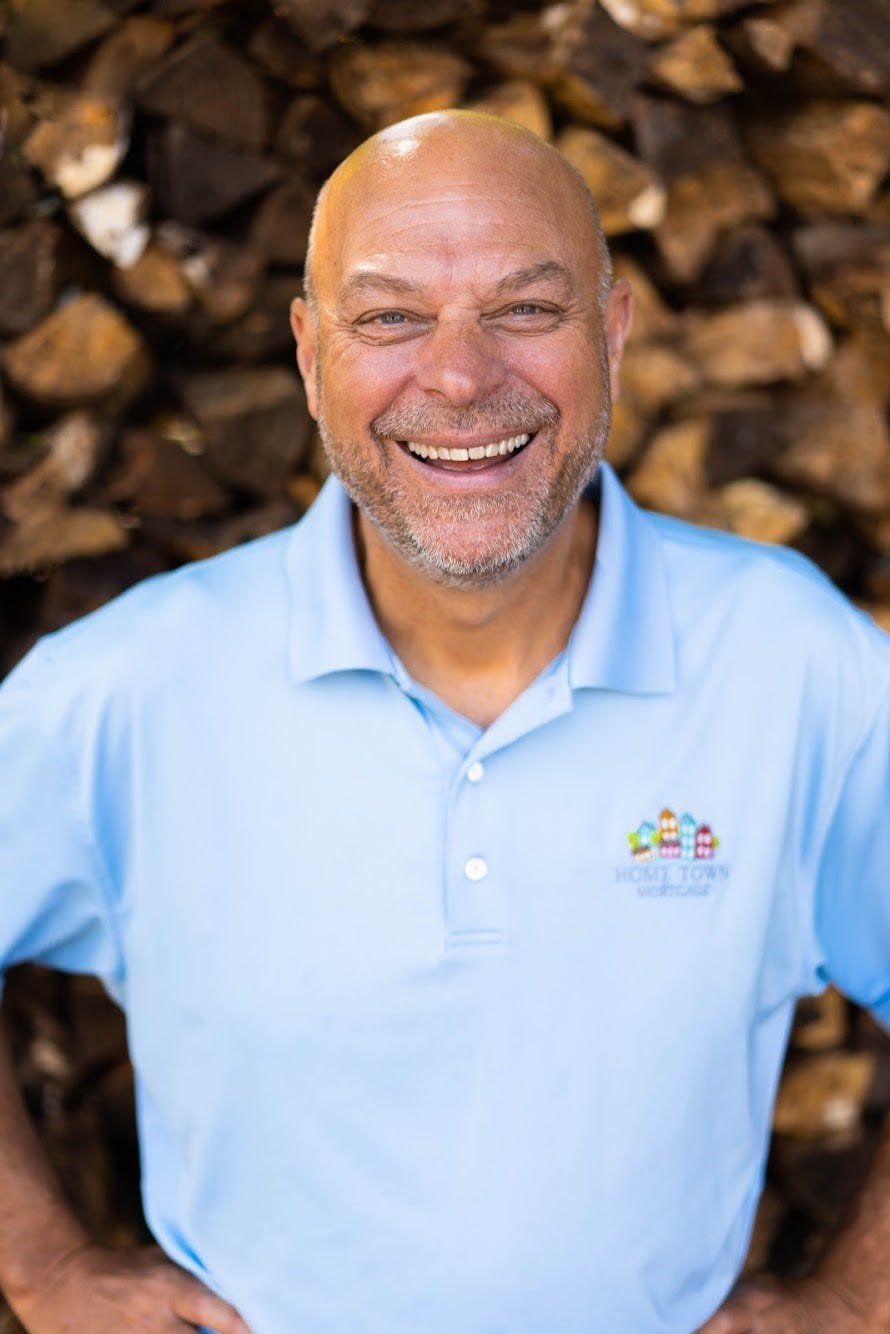 A smiling man with a bald head and gray beard wearing a light blue polo shirt with a colorful logo, standing in front of a background of stacked firewood.