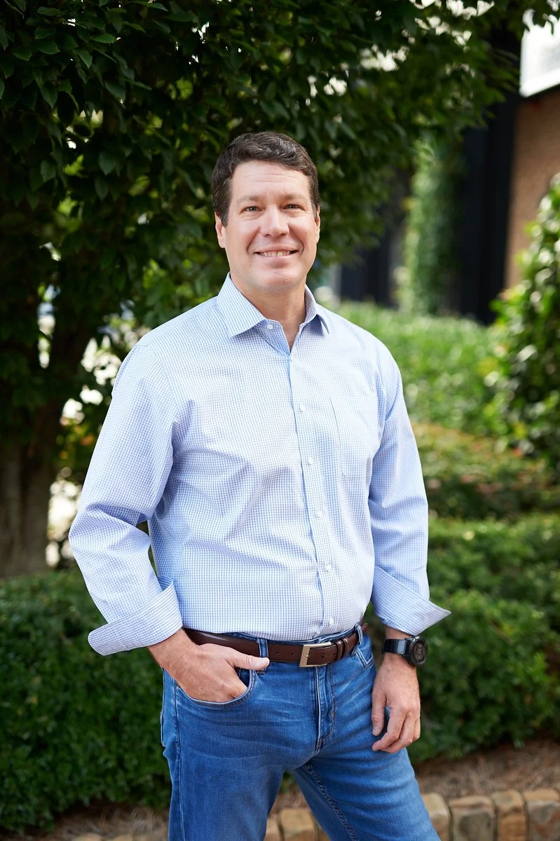 A man standing outdoors in front of green bushes, wearing a light blue button-up shirt, blue jeans, and a watch, smiling at the camera.