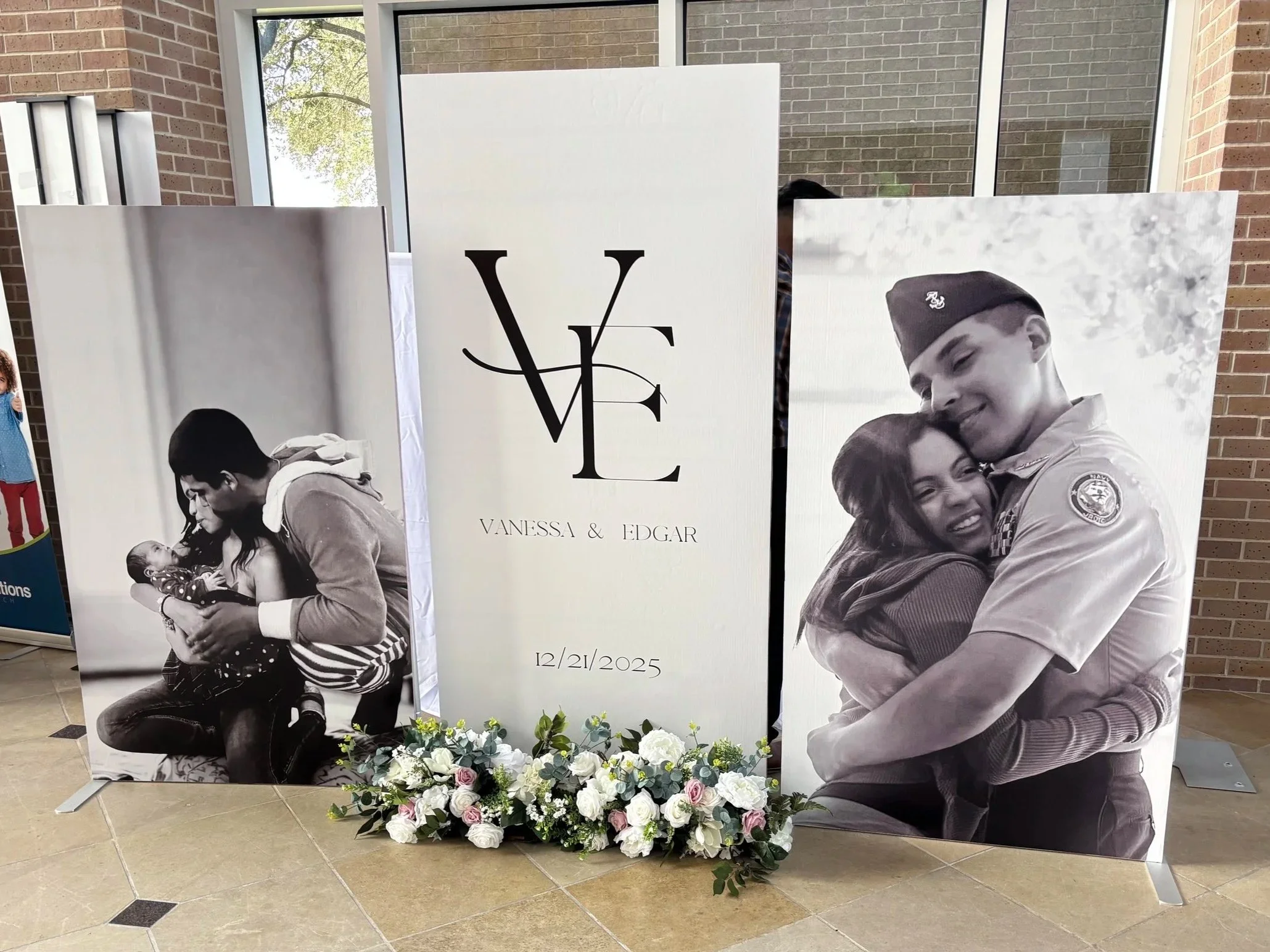 Wedding display with black-and-white photographs featuring a woman and a man in military uniform, and a central sign with the initials 'V' and 'E', the names Vanessa and Edgar, and the date 12/21/2025, surrounded by floral arrangements.