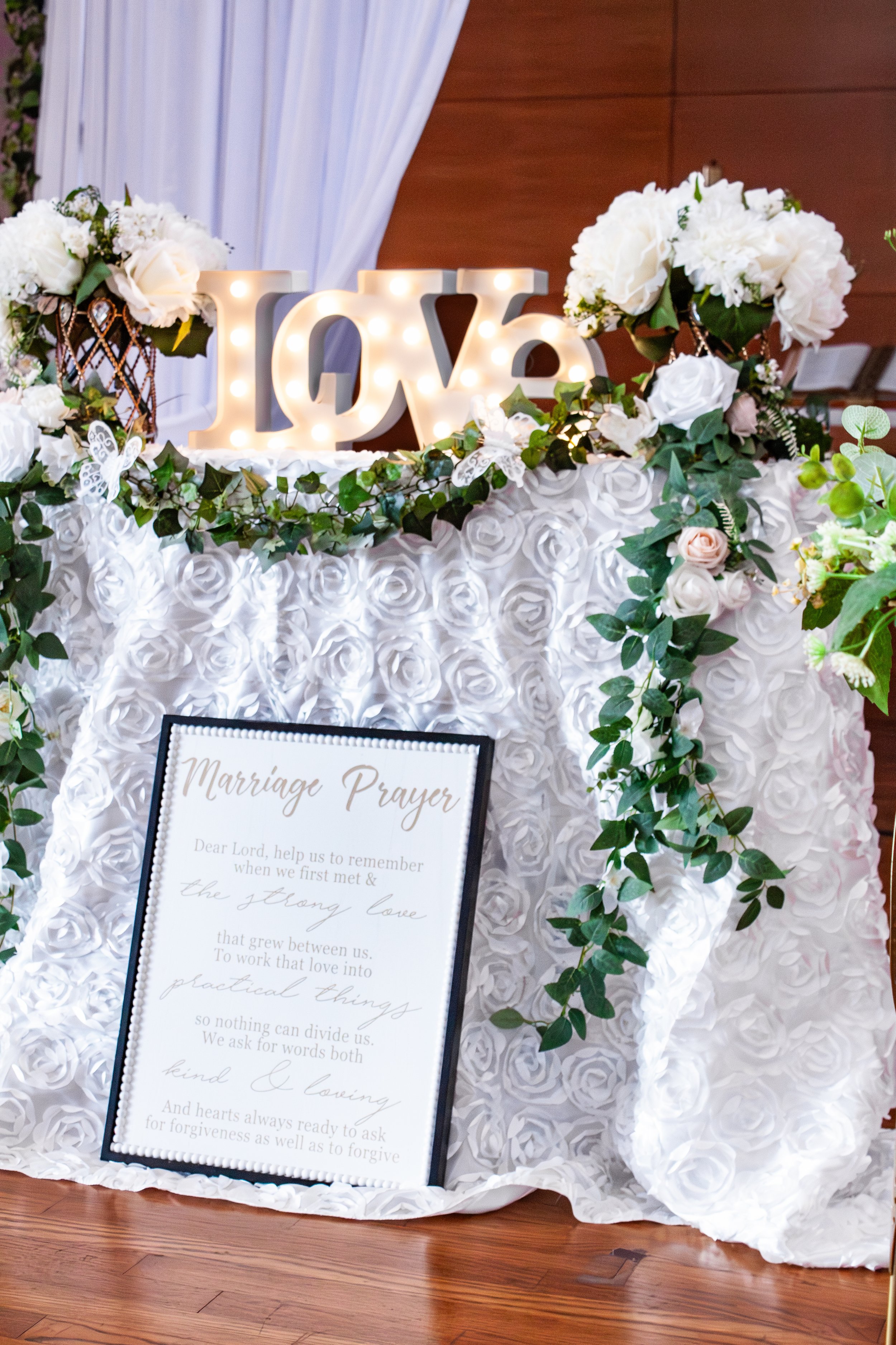 Wedding decoration with illuminated love sign, floral arrangements, and a marriage prayer sign on a white floral tablecloth.