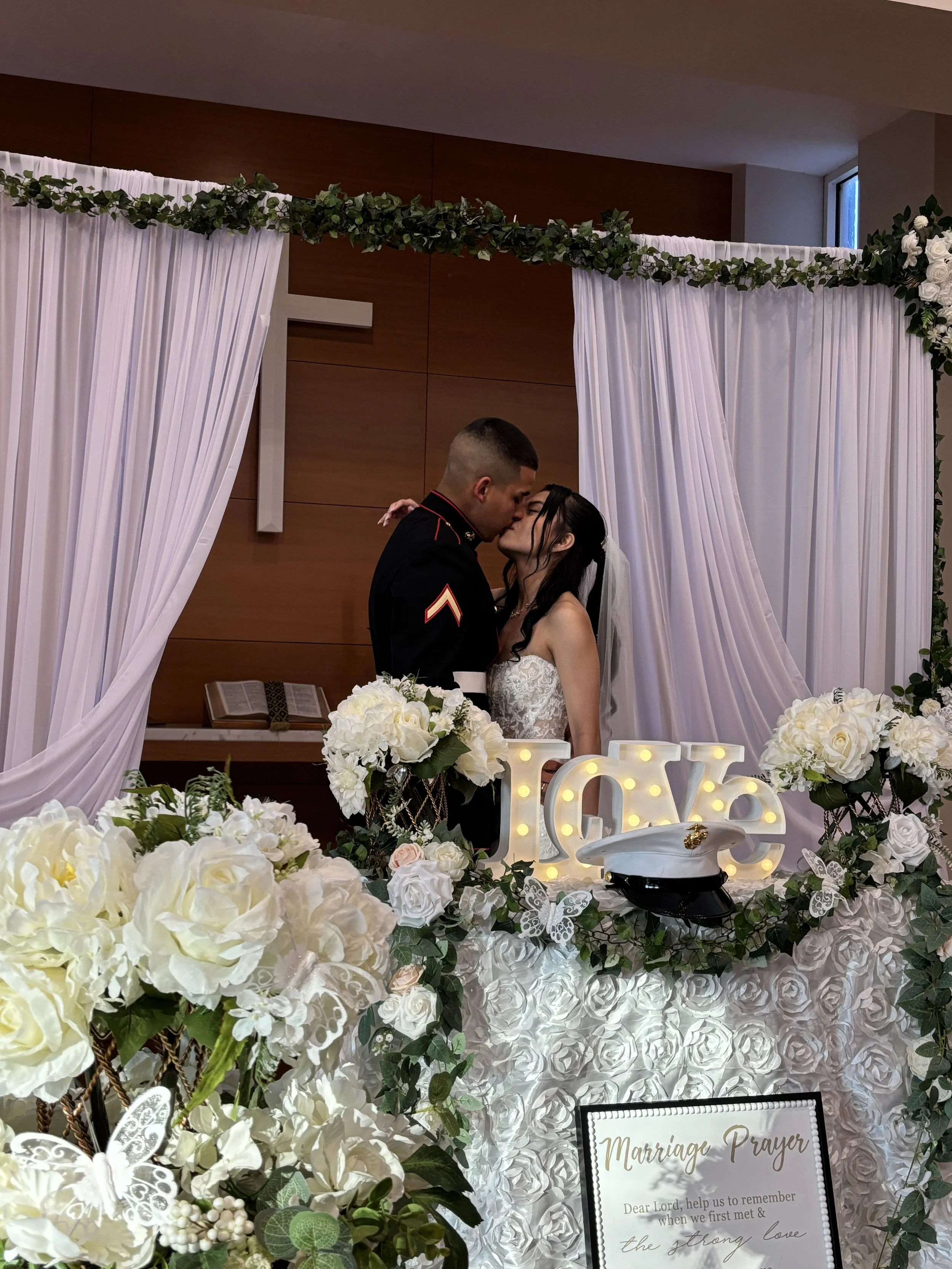 A couple kiss during their wedding ceremony, with the groom in a Marine uniform and the bride in a white wedding dress, surrounded by floral decorations and a illuminated love sign.