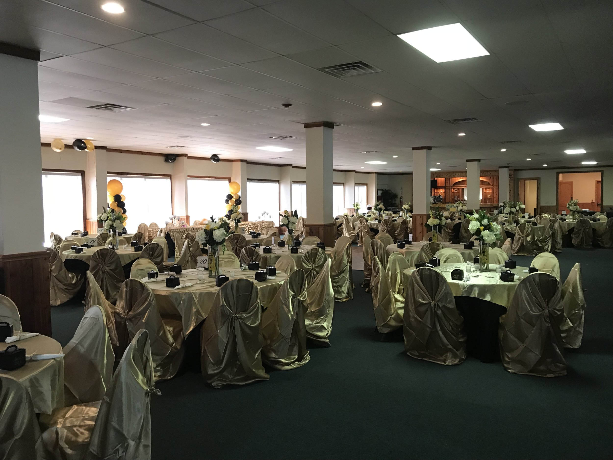 Arranged banquet hall with round tables covered in gold tablecloths, decorated with tall flower centerpieces, gold chairs with fabric sashes, and black boxes at each place setting, illuminated by overhead lighting and decorated with balloons.