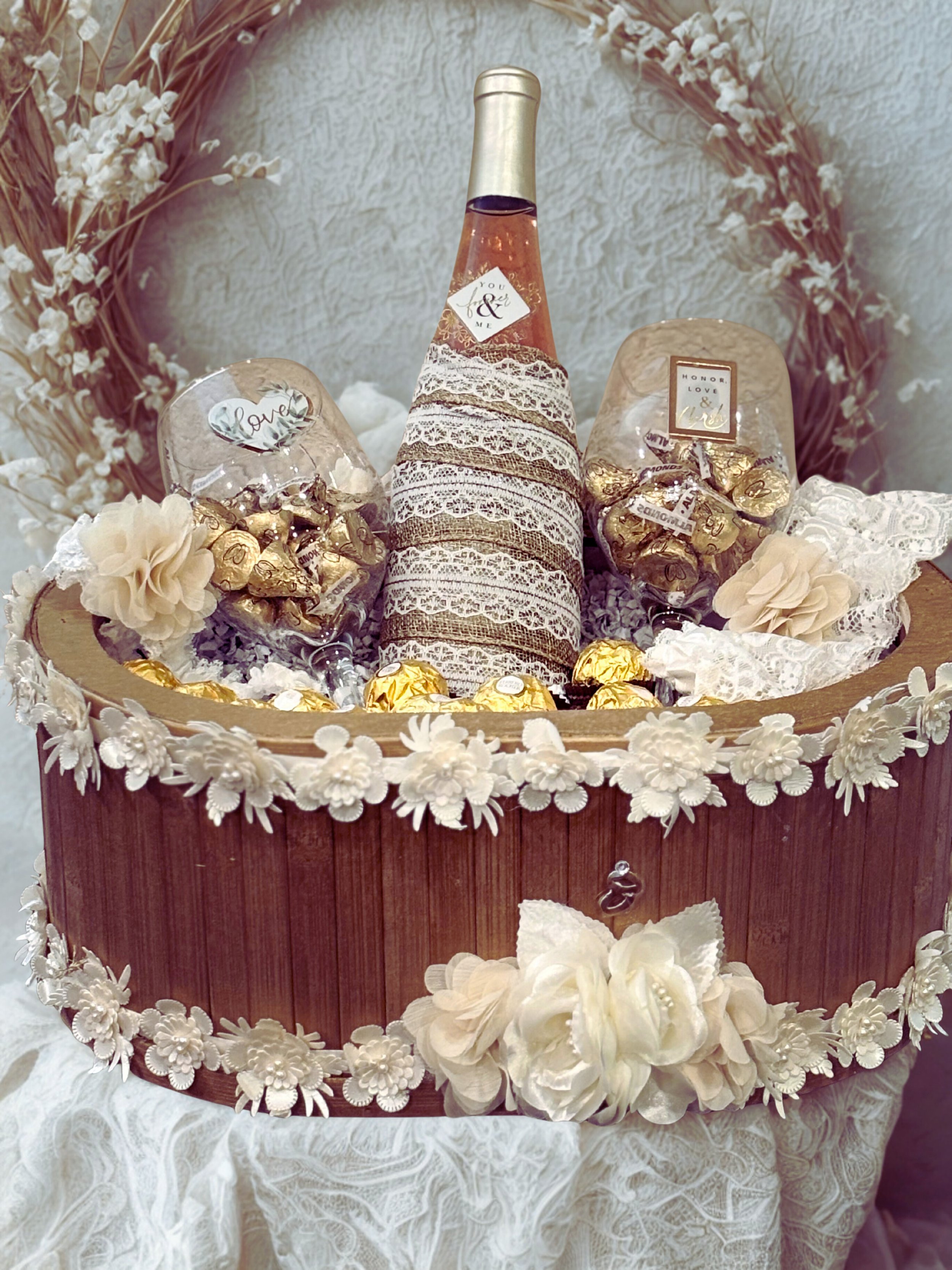Decorative gift basket with a bottle of rosé wine wrapped in lace, surrounded by chocolates and artificial flowers, on a lace-covered table with a floral wreath backdrop.