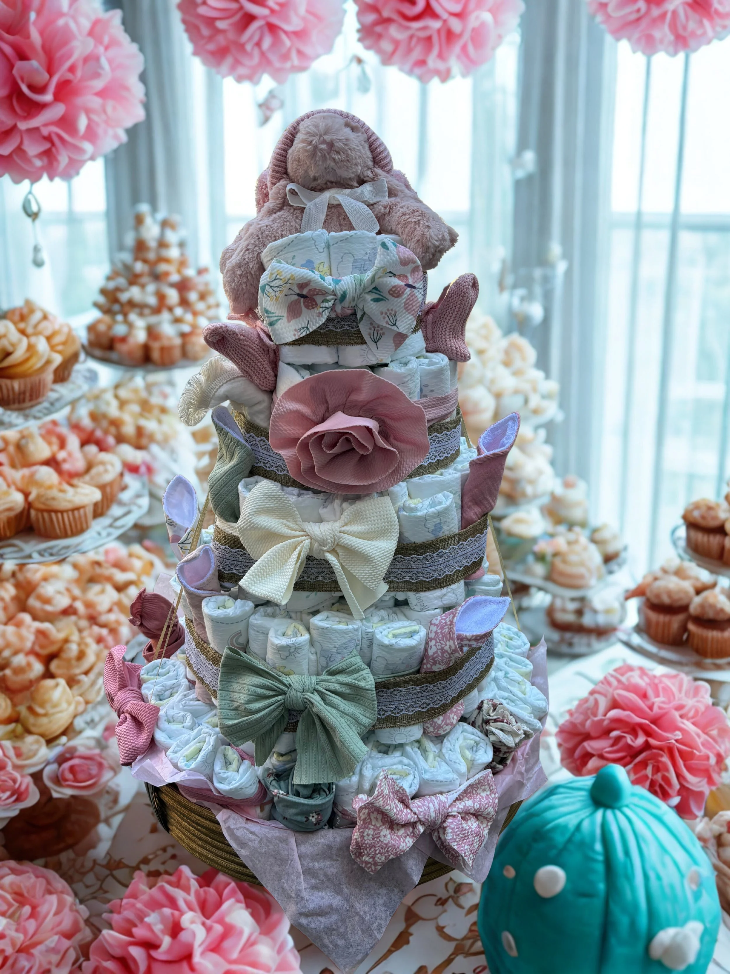 Decorative diaper cake topped with a teddy bear, surrounded by pink, white, and purple bows, lace, and fabric flowers, with cupcakes and pink flowers in the background.