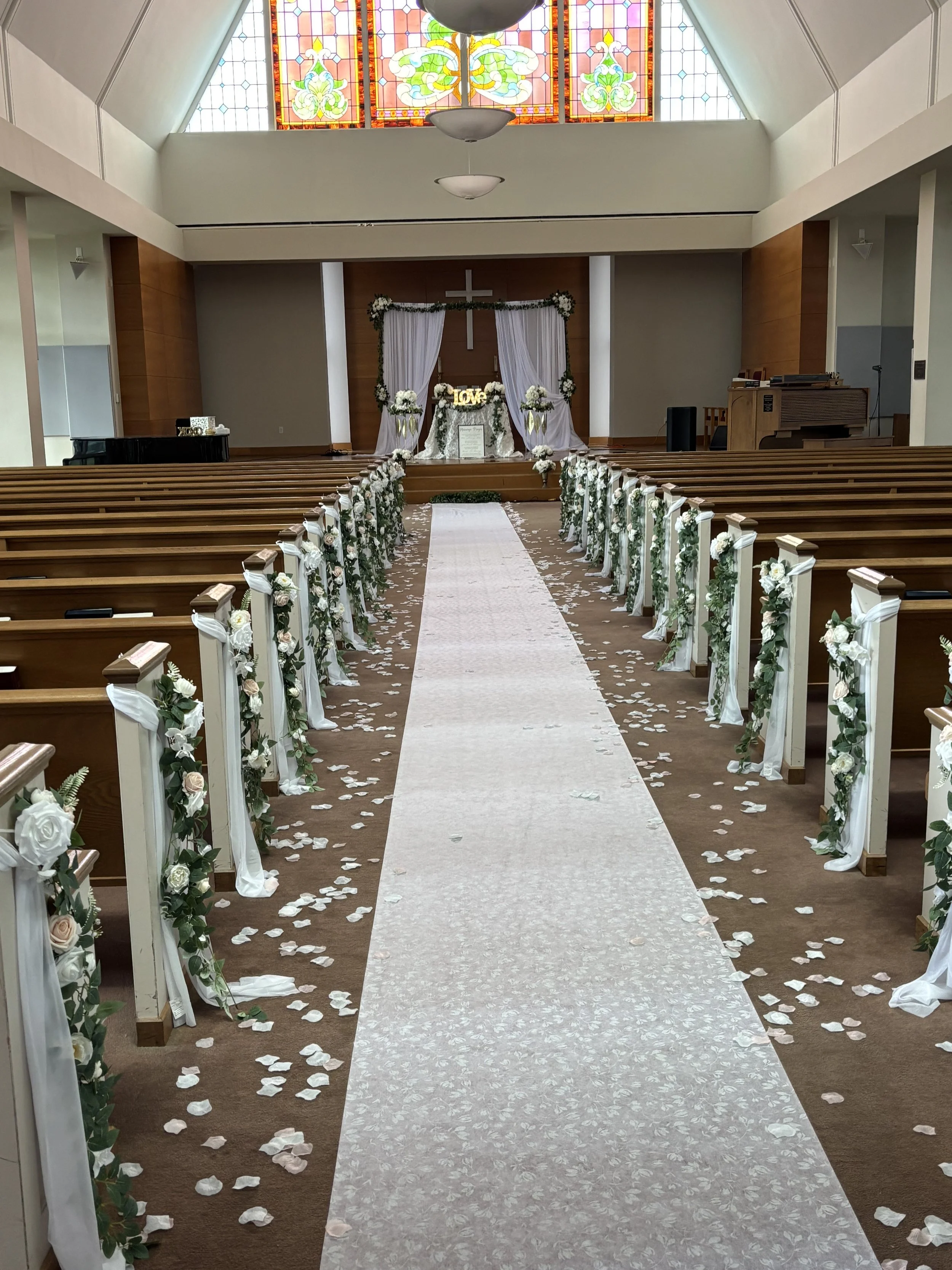 A decorated church aisle for a wedding with white flower arrangements on white pillars along the aisle, white confetti on the floor, a white aisle runner, and a decorated altar with white drapes, flowers, and a cross in the background.