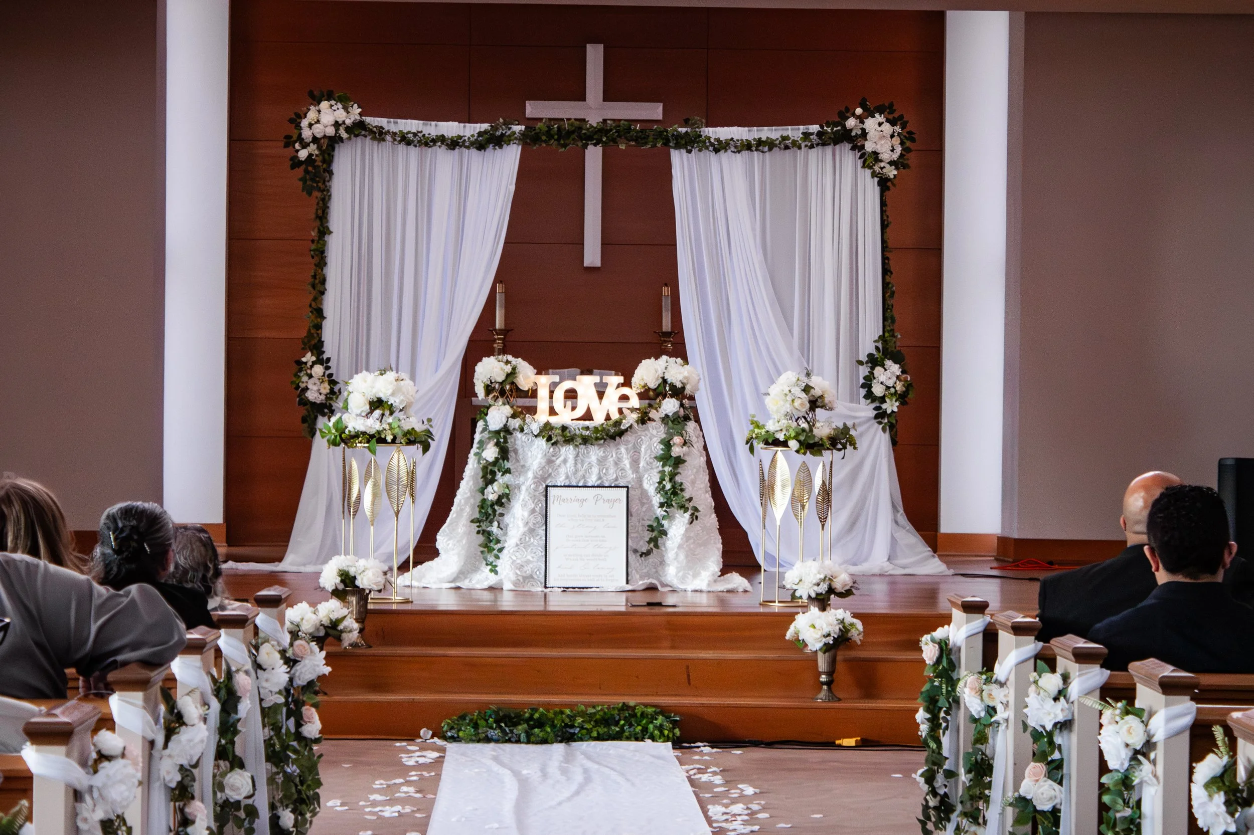 Wedding altar decorated with white flowers, draped white curtains, and a cross in the background. Guests seated on either side of an aisle with white flower decorations.