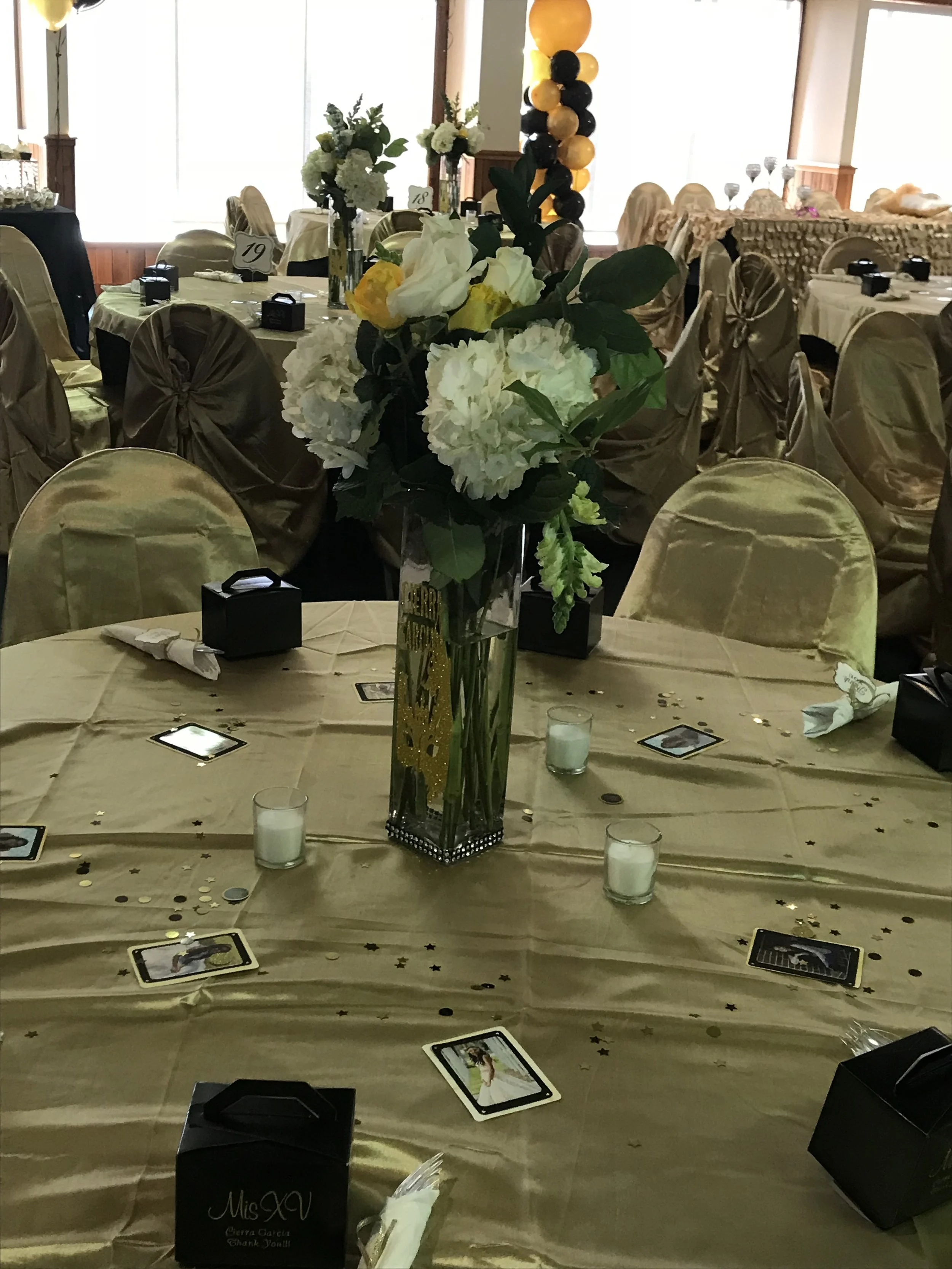 A banquet table decorated with a large bouquet of white, yellow, and green flowers in a tall glass vase, surrounded by small candles, scattered gold and black star-shaped confetti, and small black boxes, set in a decorated event hall with beige chair