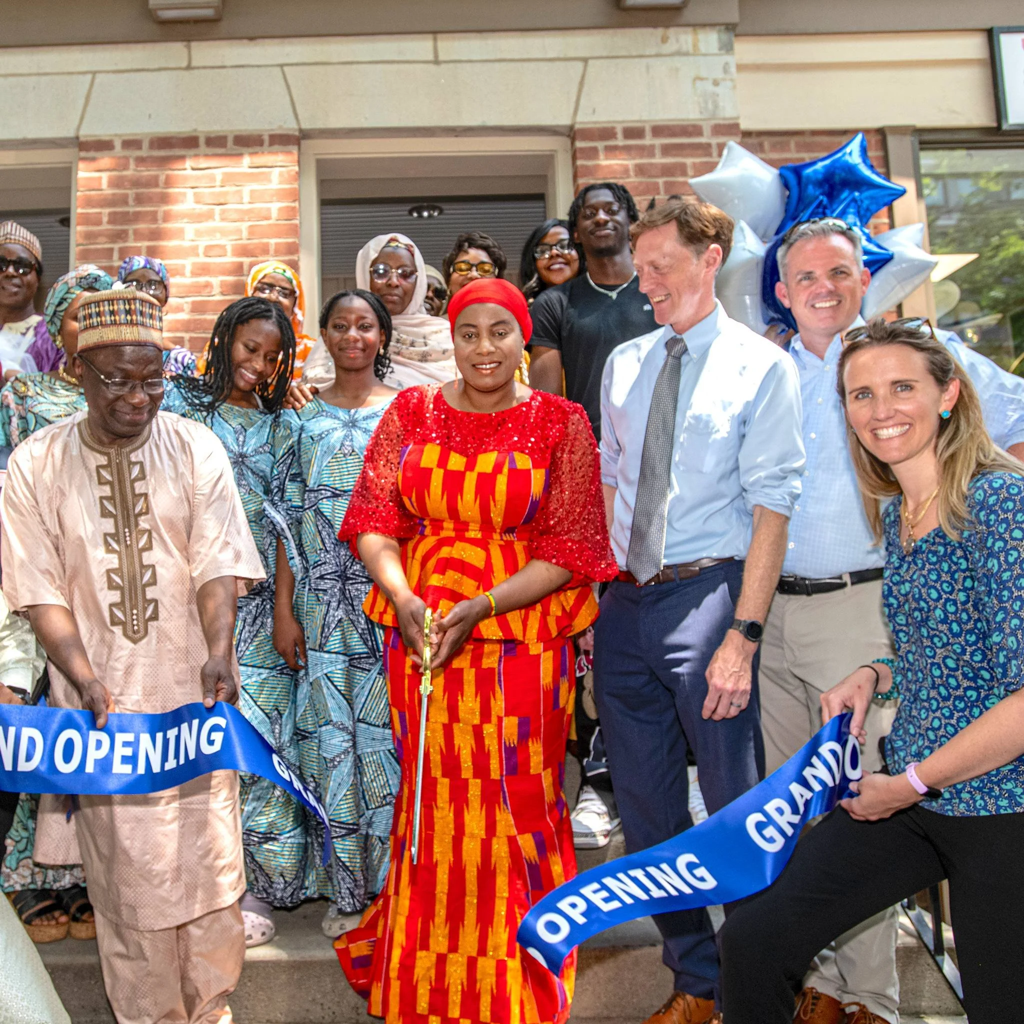 Group of people celebrating a grand opening ceremony outside a building, with a woman in traditional African attire cutting a ribbon, surrounded by smiling individuals, including men and women of diverse ethnic backgrounds.