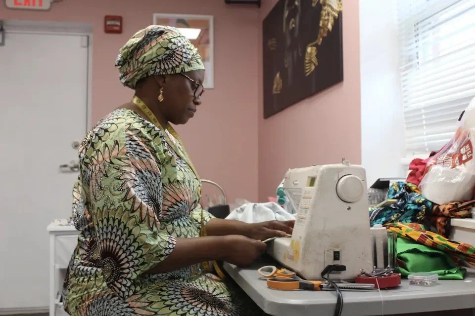 A woman wearing a colorful headwrap and dress sits at a sewing machine working on fabric, surrounded by sewing supplies and colorful fabric in a room with pink walls and a window.