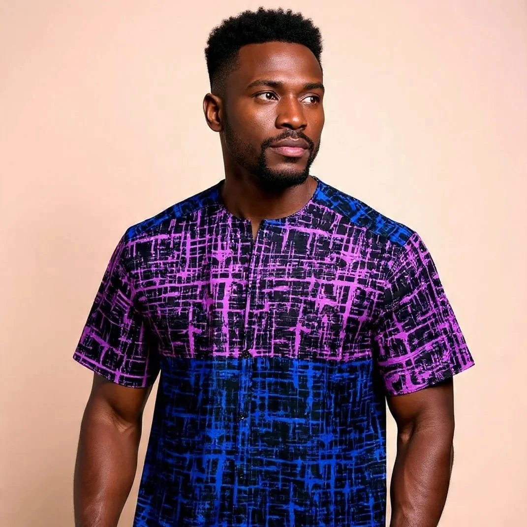 A man with dark skin and short curly hair wearing a colorful patterned shirt with purple, pink, and blue geometric designs on a light background.