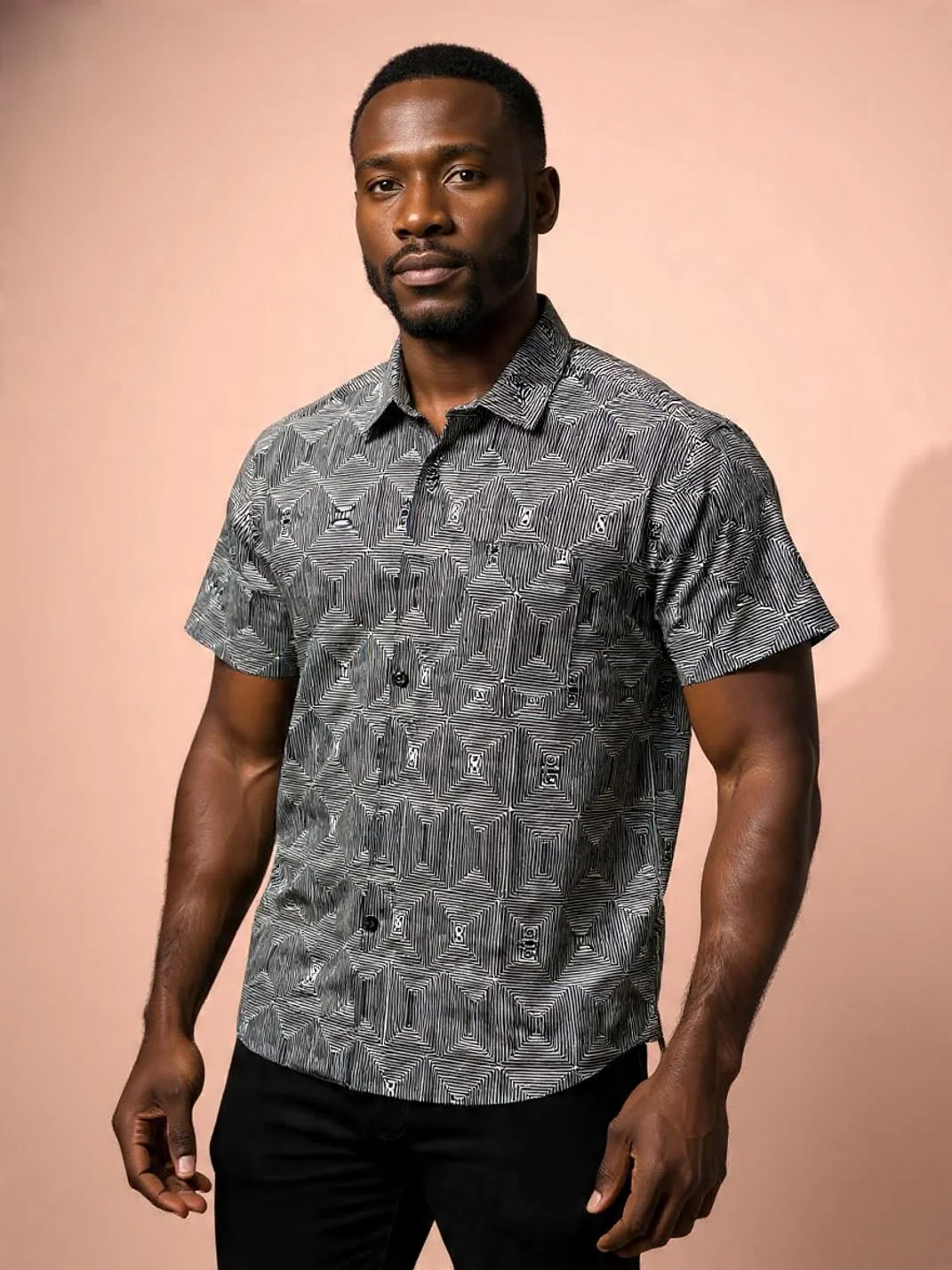 A man with dark skin, short black hair, and a beard, wearing a black and white geometric patterned short-sleeve shirt, standing against a plain light pink background.