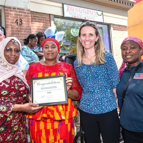 Four women standing together outside a building, holding a framed certificate. The woman second from the left is wearing a bright red and yellow dress, holding a certificate that reads 'Congratulations ZONGOZON Yale.' The woman on the far right is we