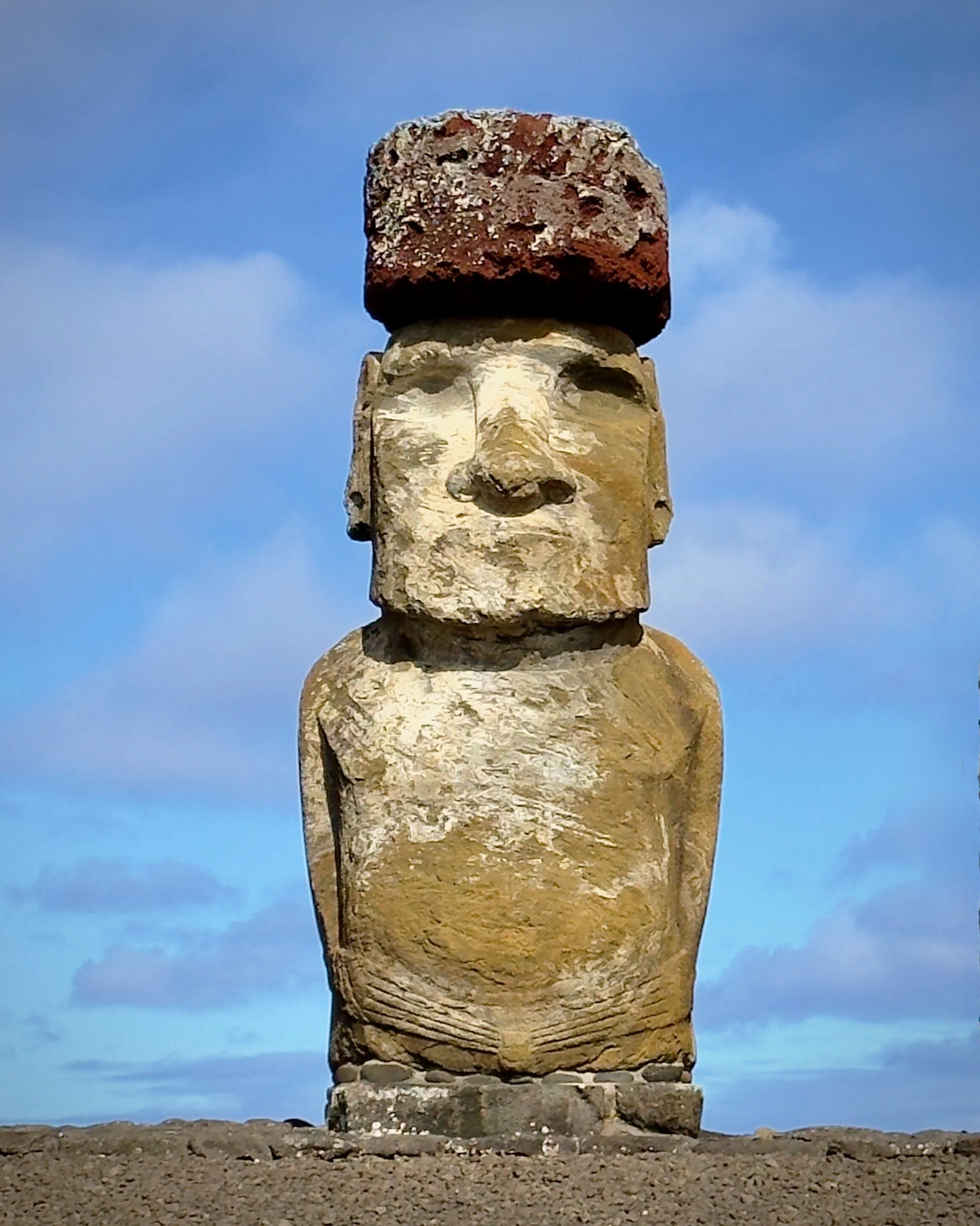 A large stone Moai statue with a red stone hat, standing on a plain with a cloudy sky in the background.