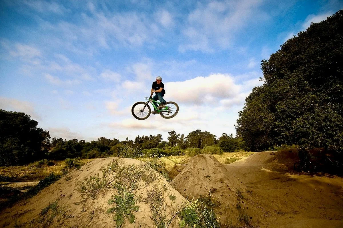Person in black shirt and helmet riding a green dirt bike mid-air over dirt mounds on a dirt trail with trees and blue sky in the background.