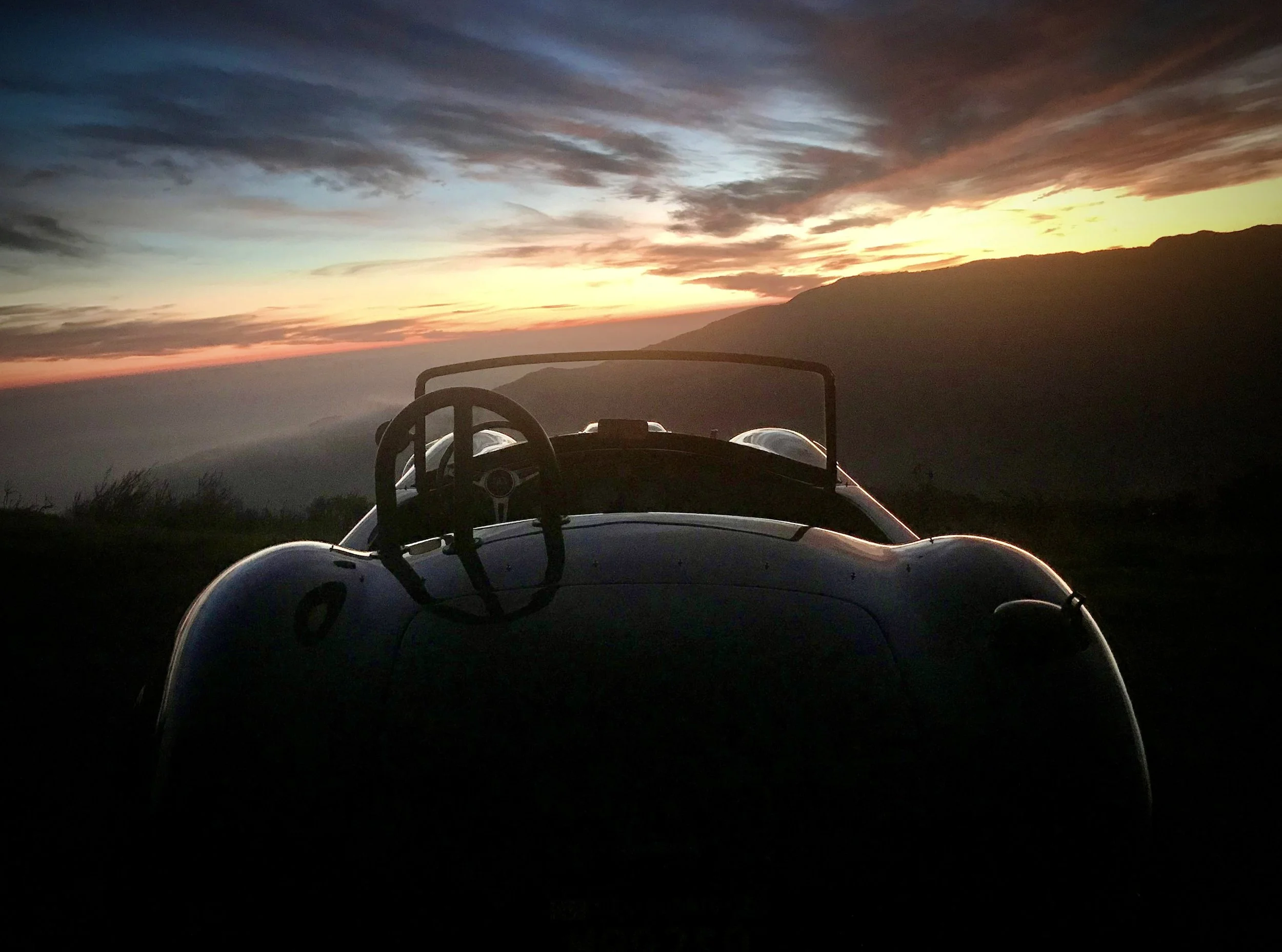 Silhouette of a vintage race car with a steering wheel and windshield, set against a scenic sunset with mountains in the background.