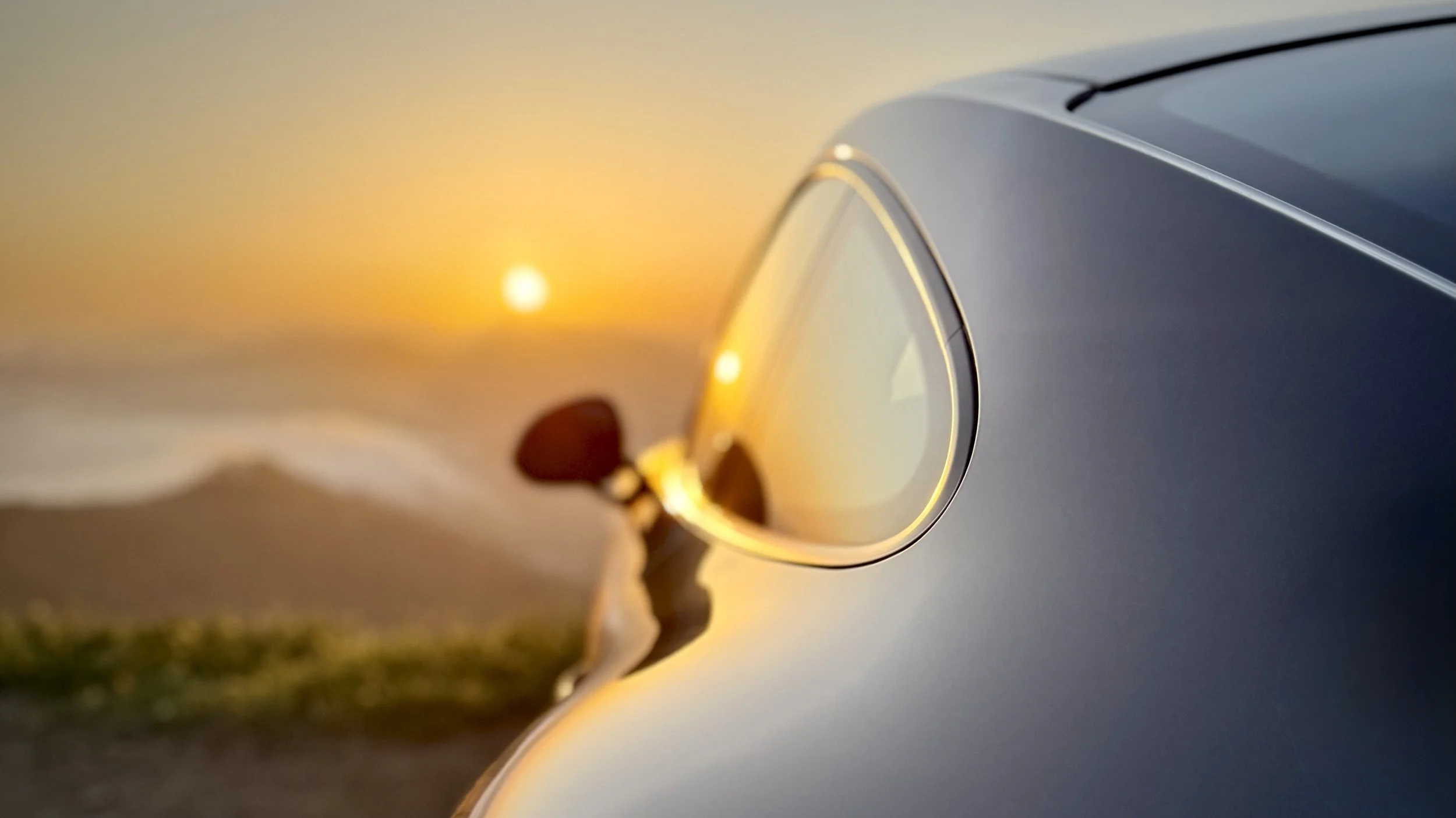 Close-up of a silver car's side mirror, reflecting the sunset with orange and yellow hues, against a blurred landscape background.
