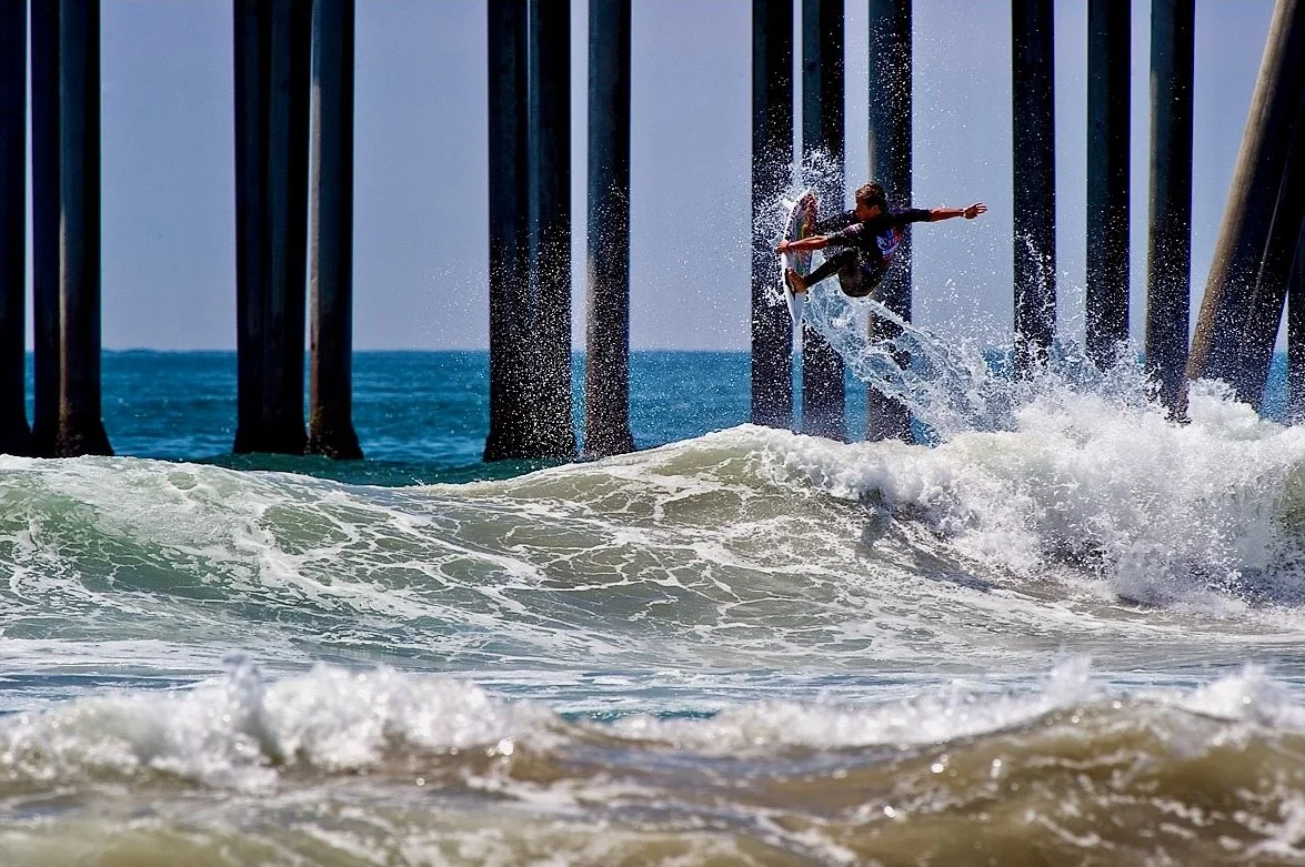 A person surfing on a wave under a pier with wooden supports.