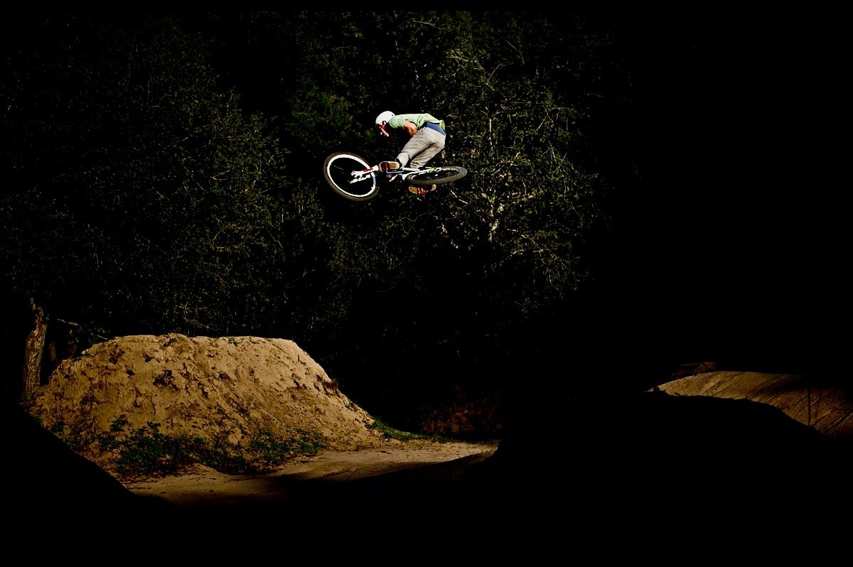 A person performing a jump on a BMX bike at night, with dirt jumps and trees in the background.