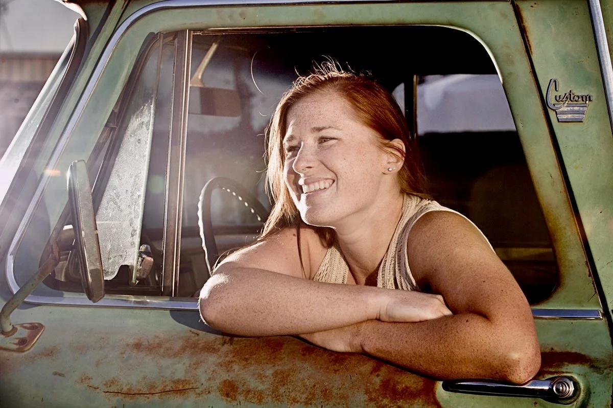 A woman with red hair and freckles smiling as she leans on the window of an old, weathered green truck.