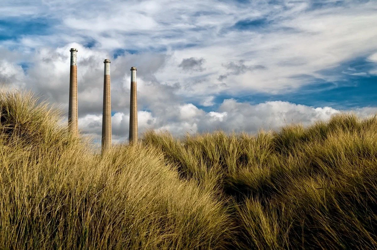 Three tall industrial smokestacks rising above grassy sand dunes with a cloudy sky in the background.
