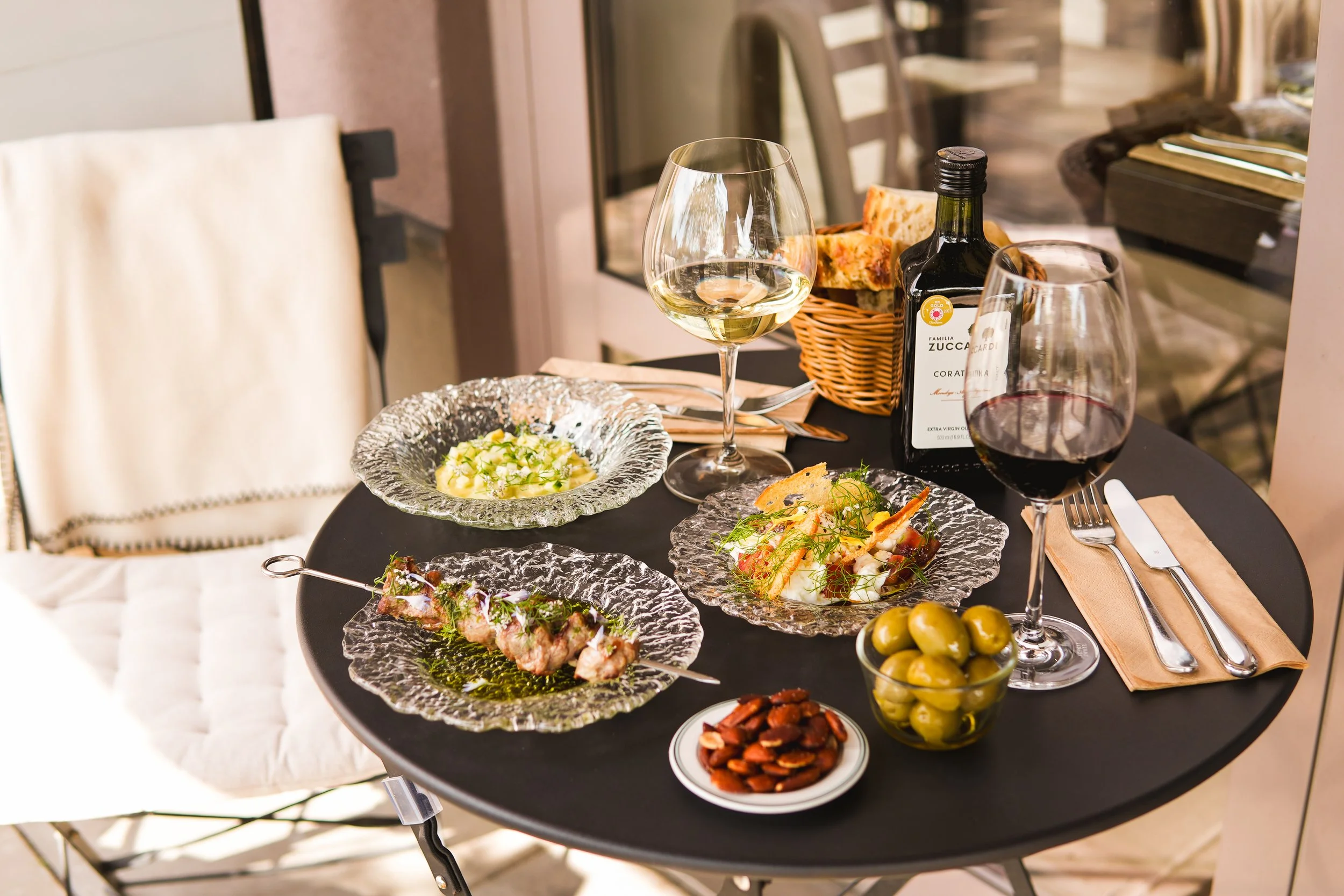 A table set for dining with glassware, plates, and dishes of food, including green olives, sliced almonds, and a skewer of meat. There is a bottle of white wine, a glass of white wine, and a glass of red wine. The table is on a balcony or patio with 