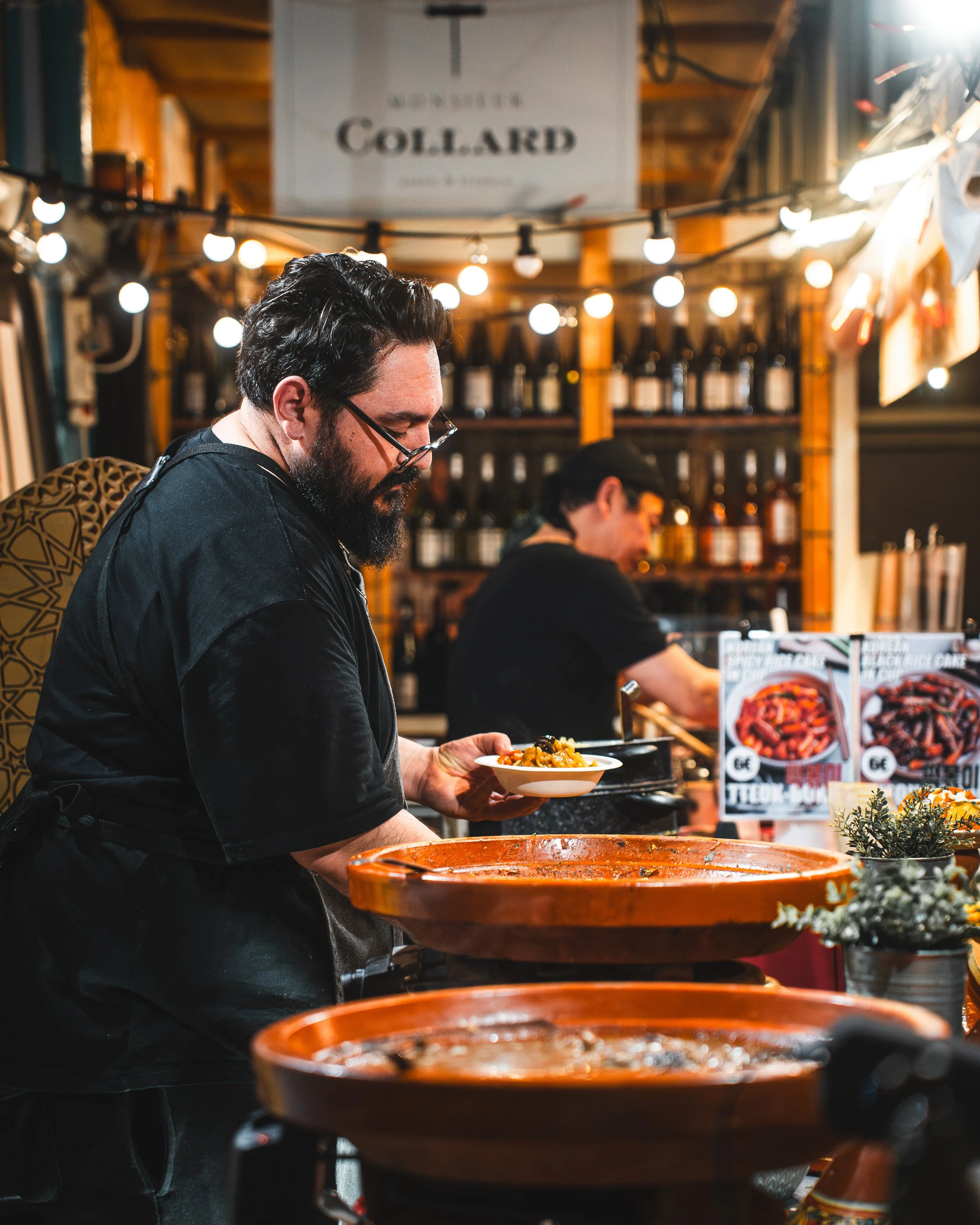 A man with glasses and a beard serving food at a restaurant or food stall decorated with string lights and a sign reading 'Collard'. He is holding a bowl of food and standing near pots of cooking ingredients or sauces, with a woman in the background 
