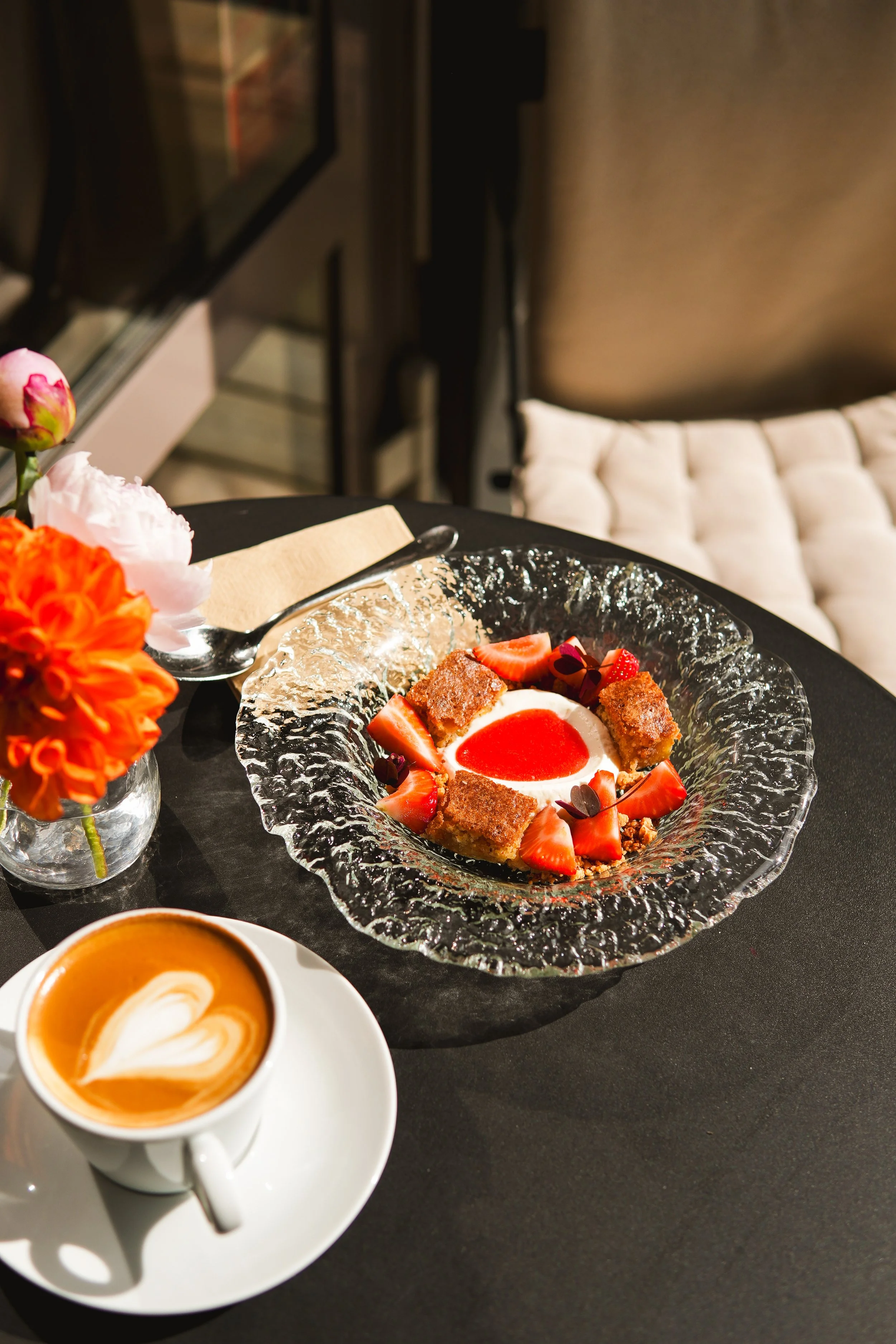 A dessert with strawberries, whipped cream, and caramelized pieces served on a textured glass plate, with a cup of latte with latte art and a small vase with orange and pink flowers on a black table.