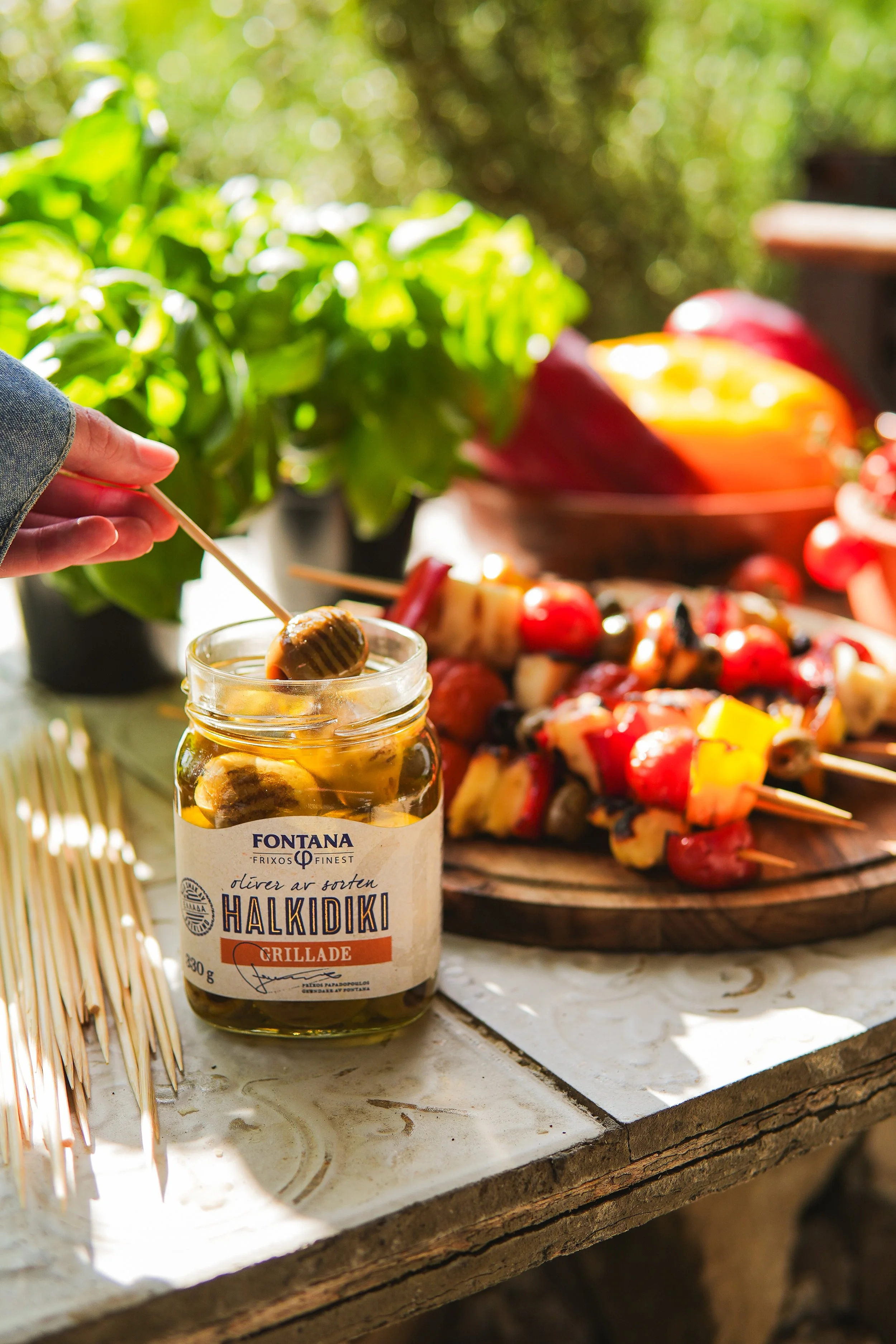 Close-up of a jar of Greek-style olive and roasted pepper spread on a table, with skewers of grilled vegetables and fruits, and a hand dipping a skewer into the jar. The background features green plants and a sunny outdoor setting.