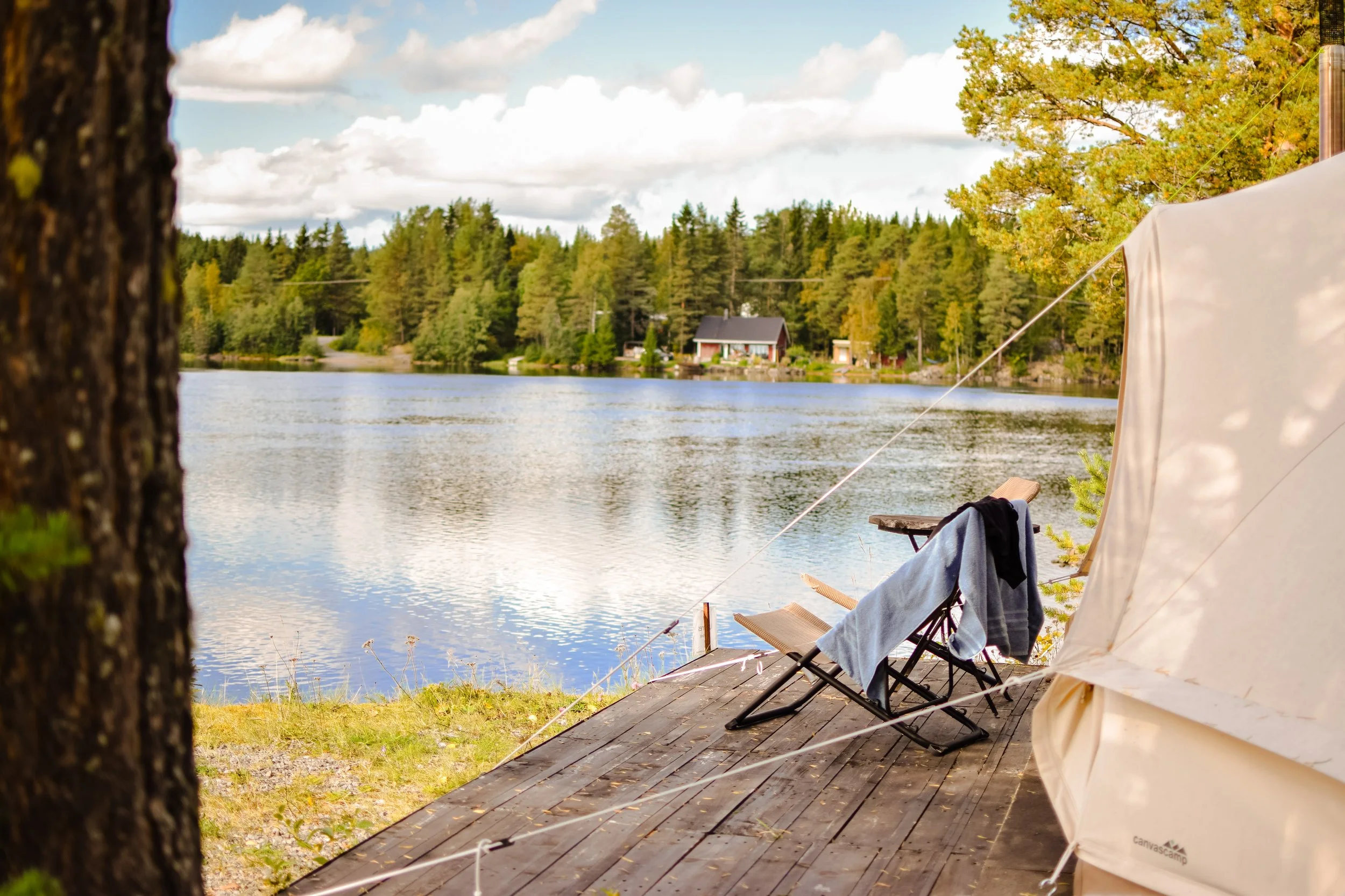 A lakeside camping scene with a tent, a chair with clothes draped over it, and trees in the background under a partly cloudy sky.