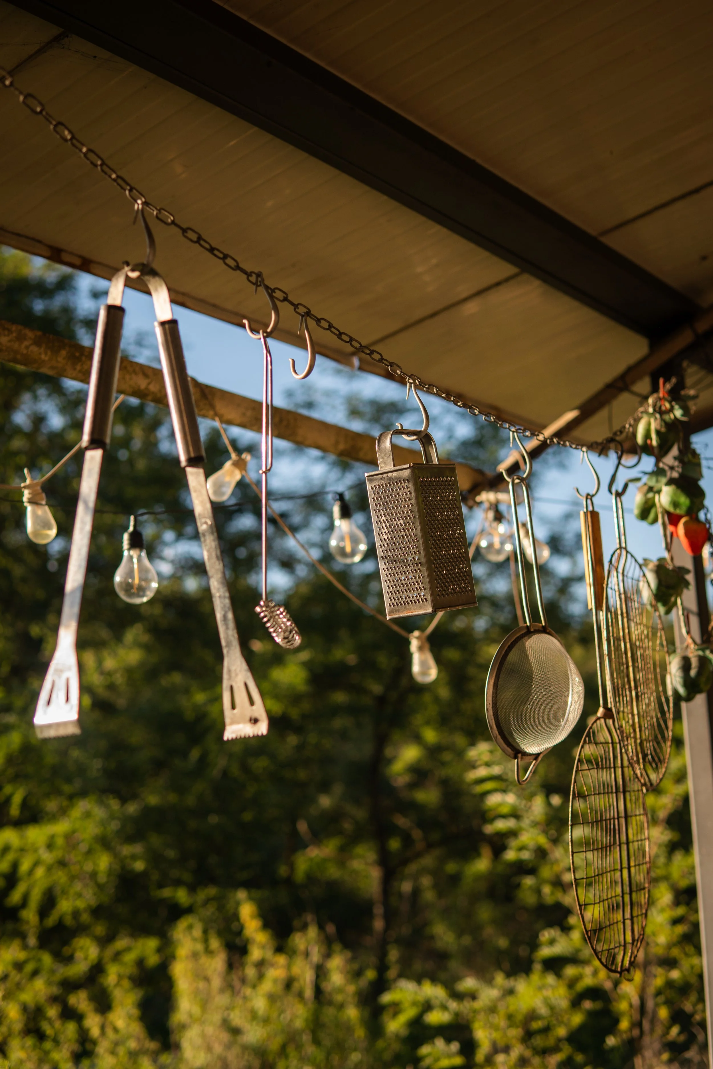 Hanging kitchen utensils and tools, including a grater, skillet, grill grate, and kitchen tongs, with string lights and outdoor trees in the background.
